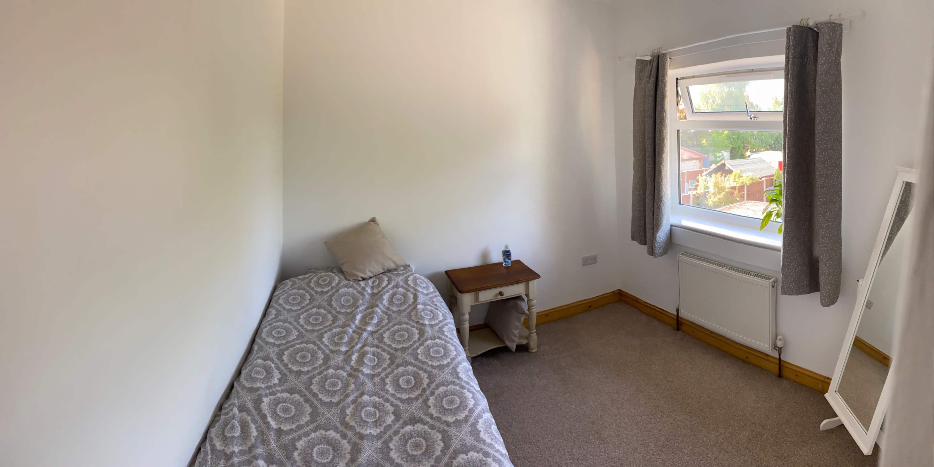 The image shows a simple bedroom with a single bed, a small bedside table, and a window overlooking a garden.