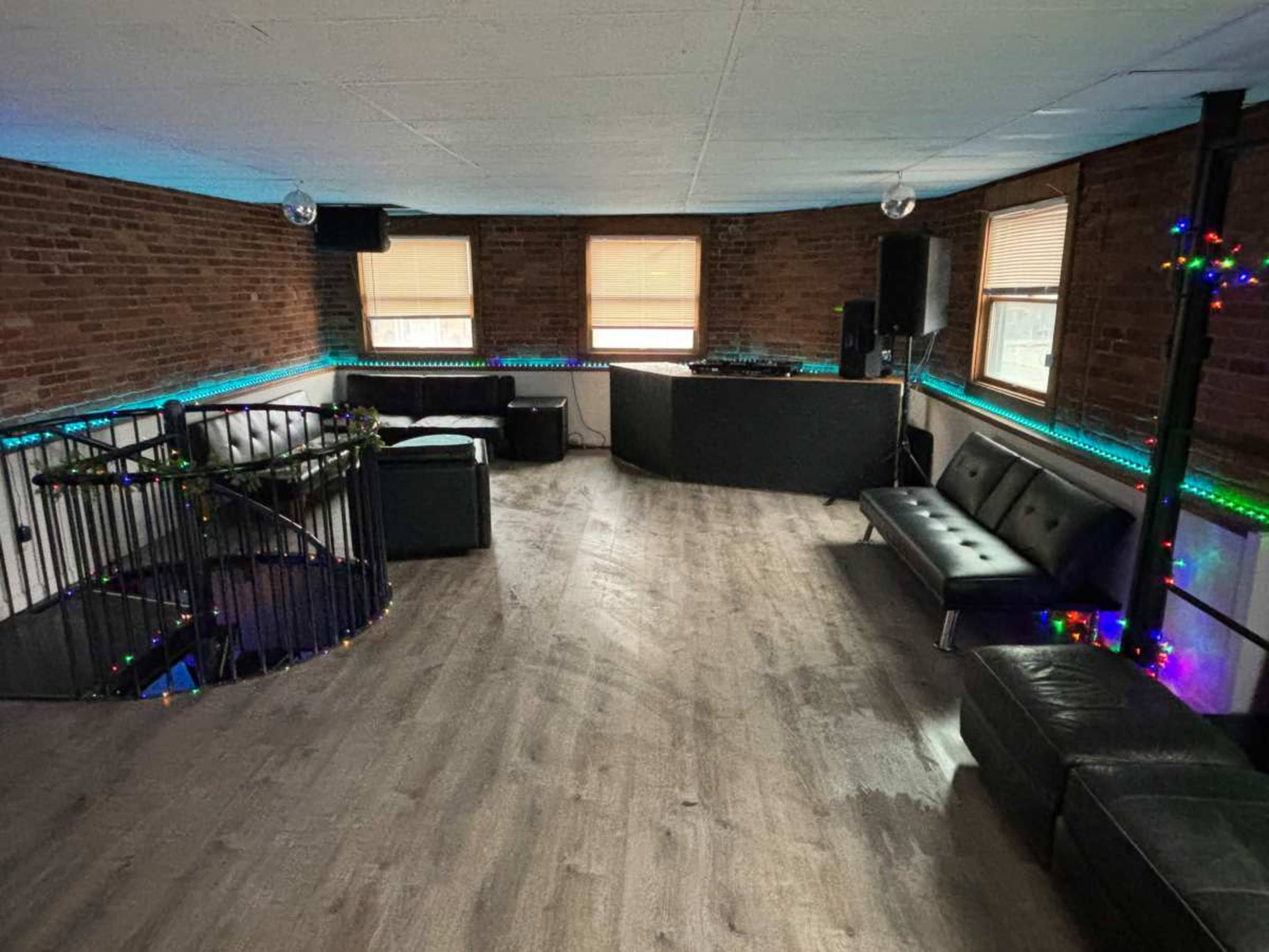 A spacious, unfinished room with exposed brick walls, a DJ setup, and seating arranged around a spiral staircase.