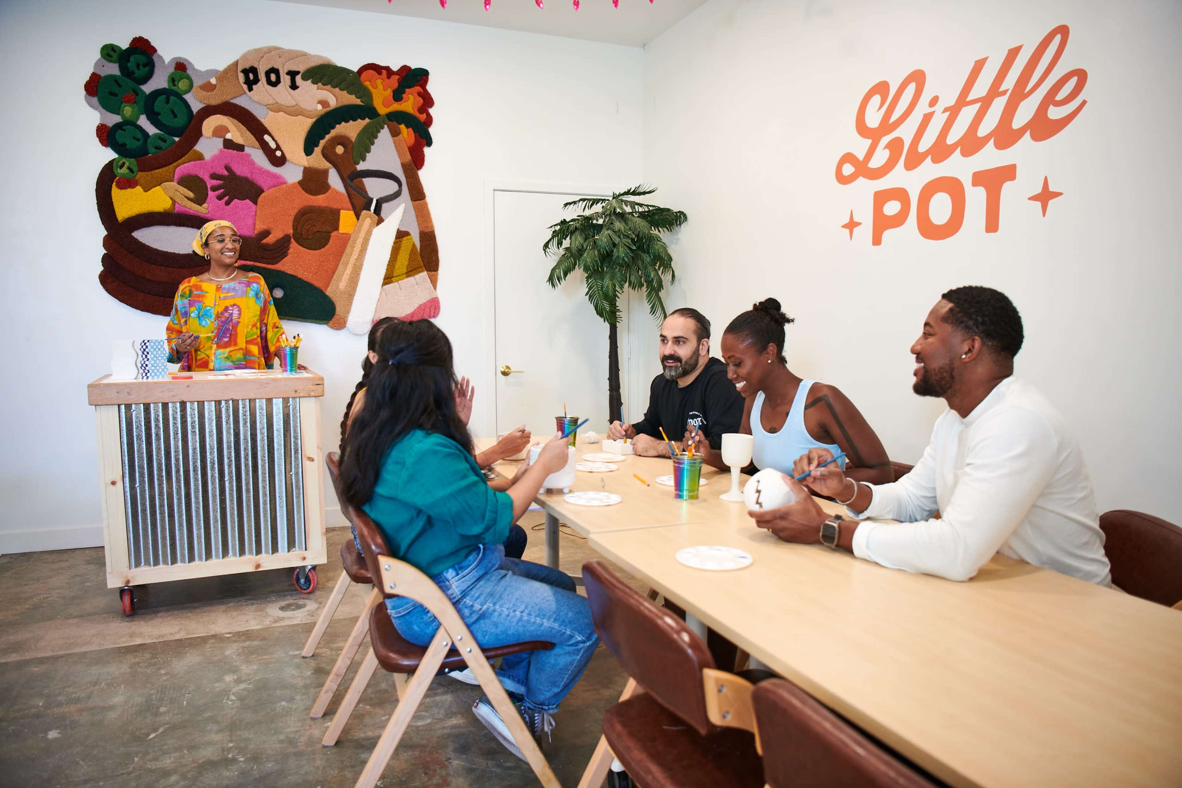 A group of five people is seated at a long table in a brightly lit room with a mural on the wall and a person standing behind a counter.