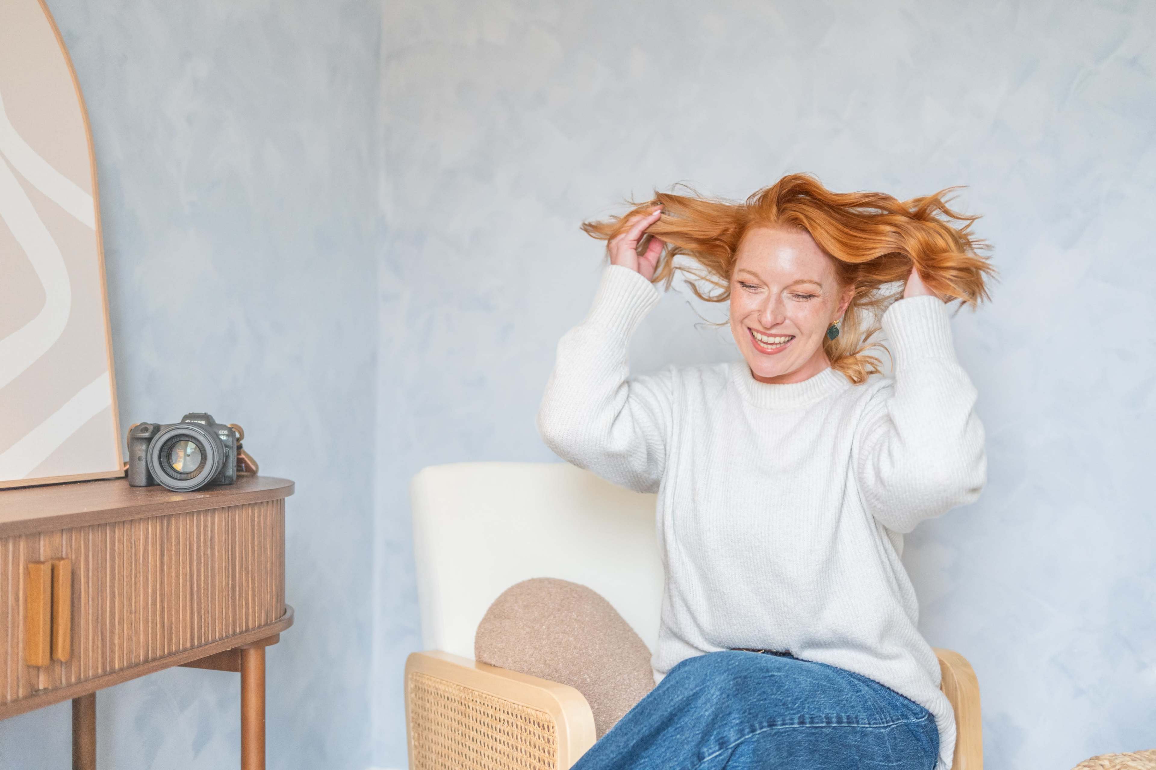 A woman with long red hair smiles while playfully tossing her hair in a well-lit room with light blue walls and a vintage camera on a nearby table.