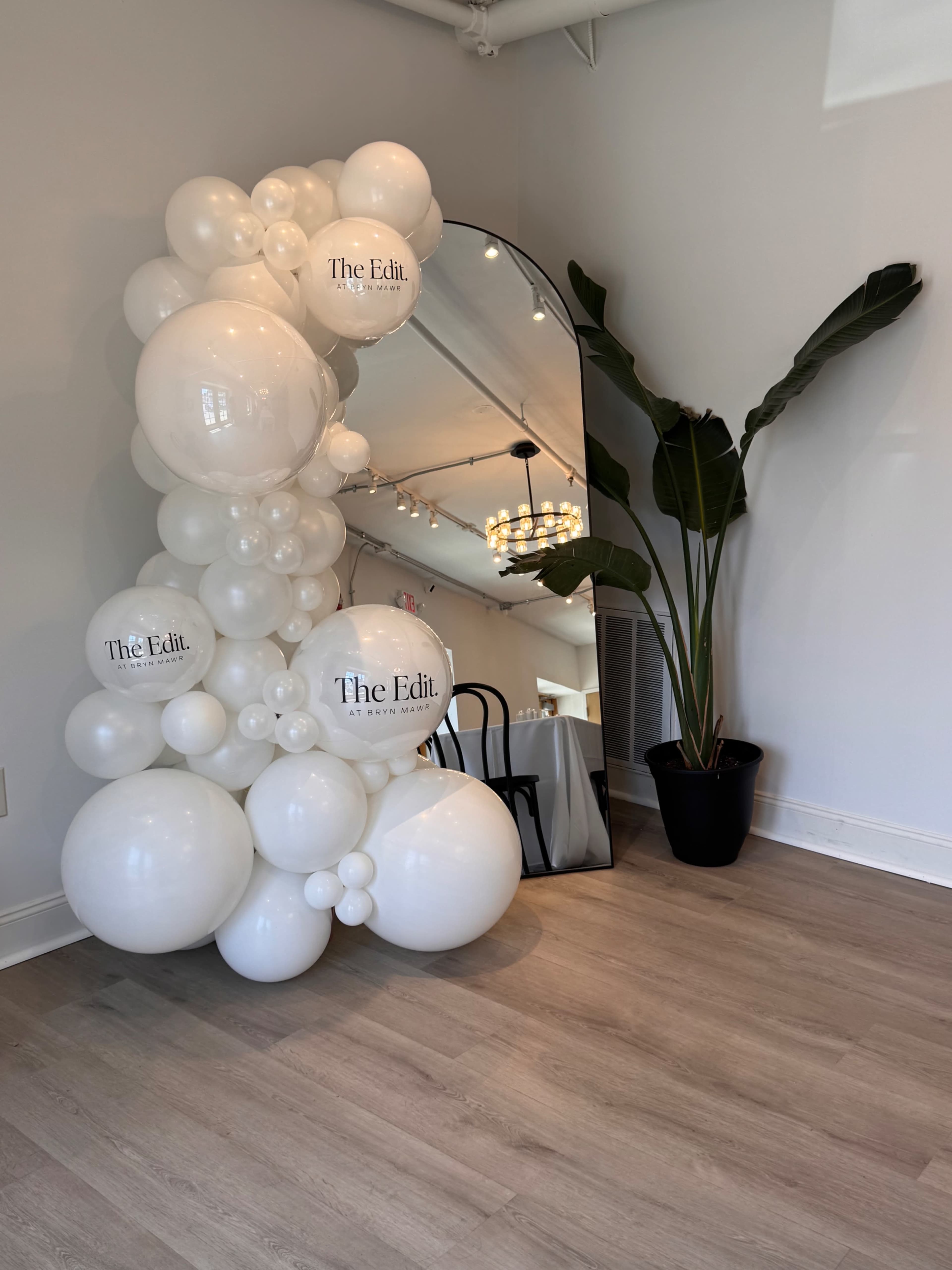 A cluster of white balloons with the text "The Edit." is arranged next to a large mirror and a tall plant in a modern interior space.