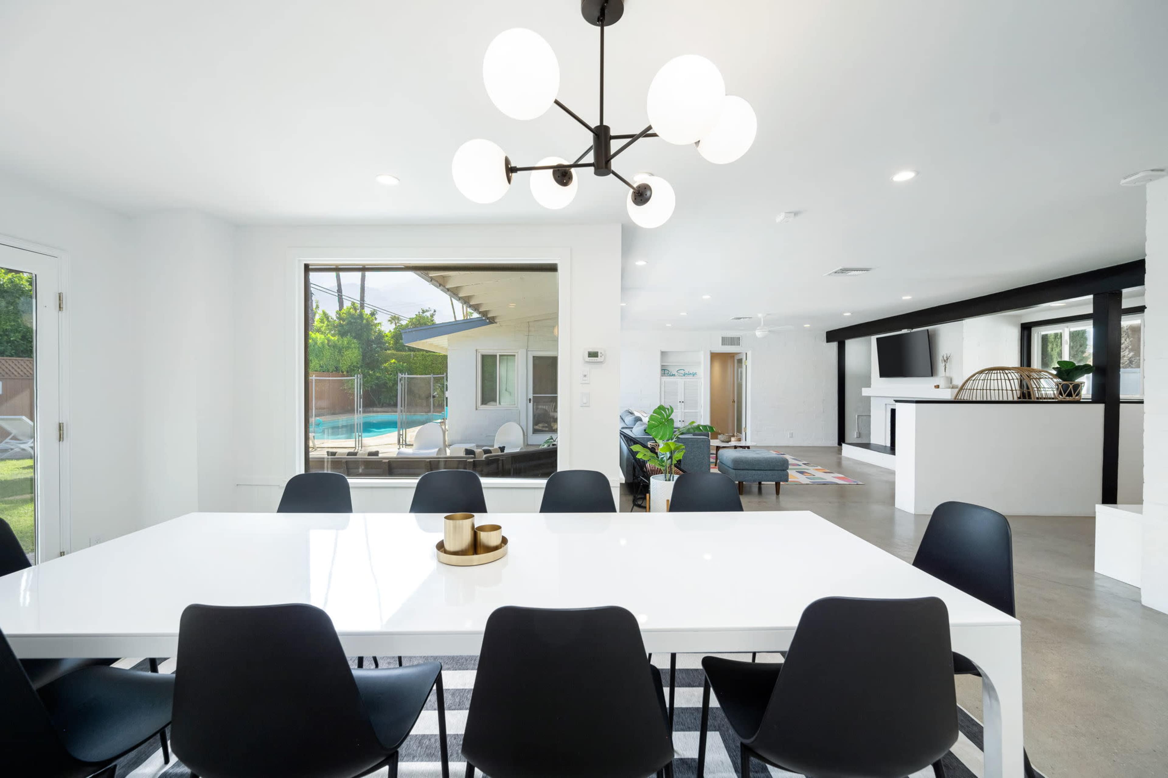 A modern dining area features a large white table surrounded by black chairs, with a view of a swimming pool and living area through large windows.