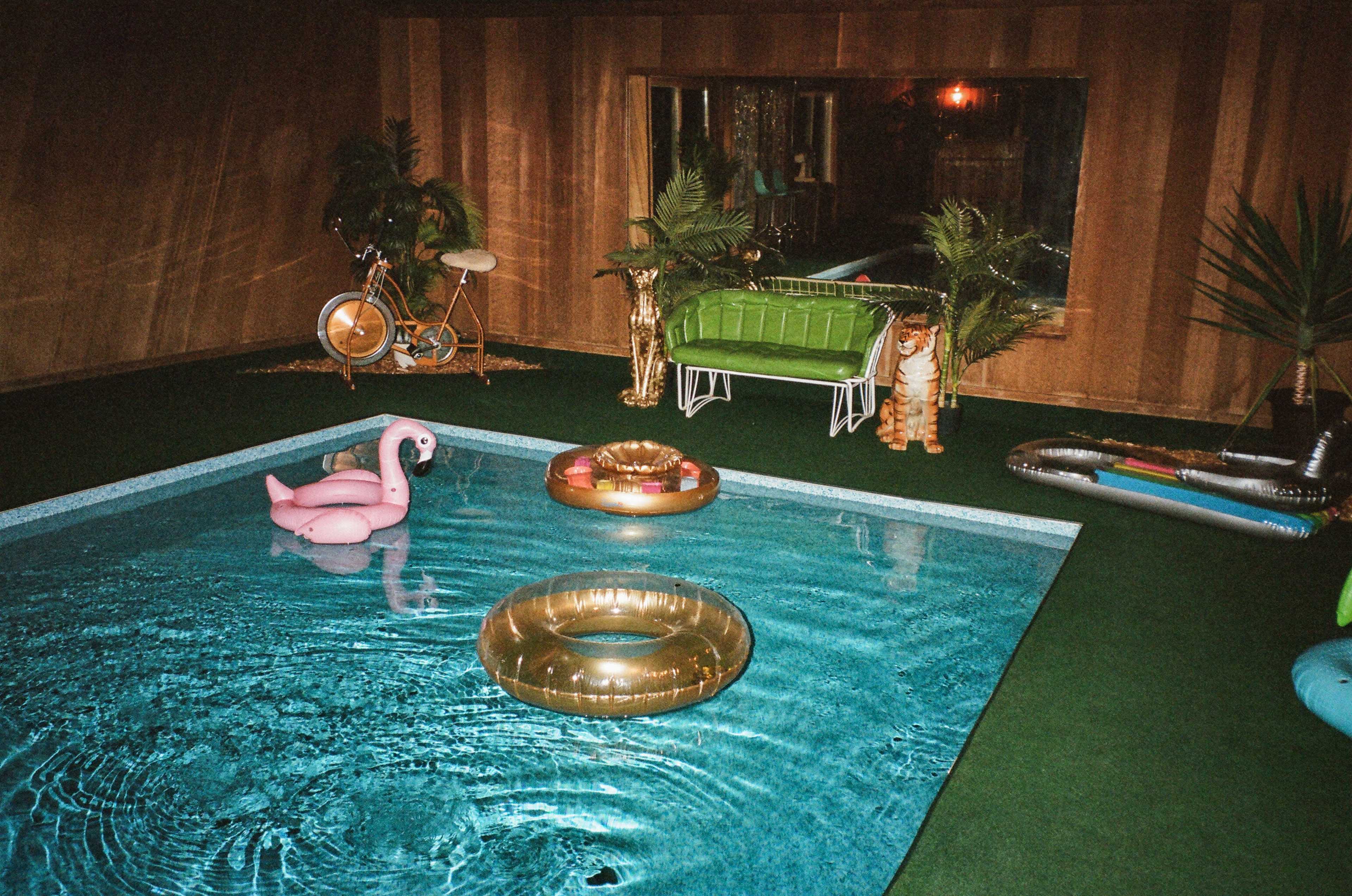 The image depicts a cozy indoor swimming pool area featuring inflatable pool floats, greenery, and retro furniture.