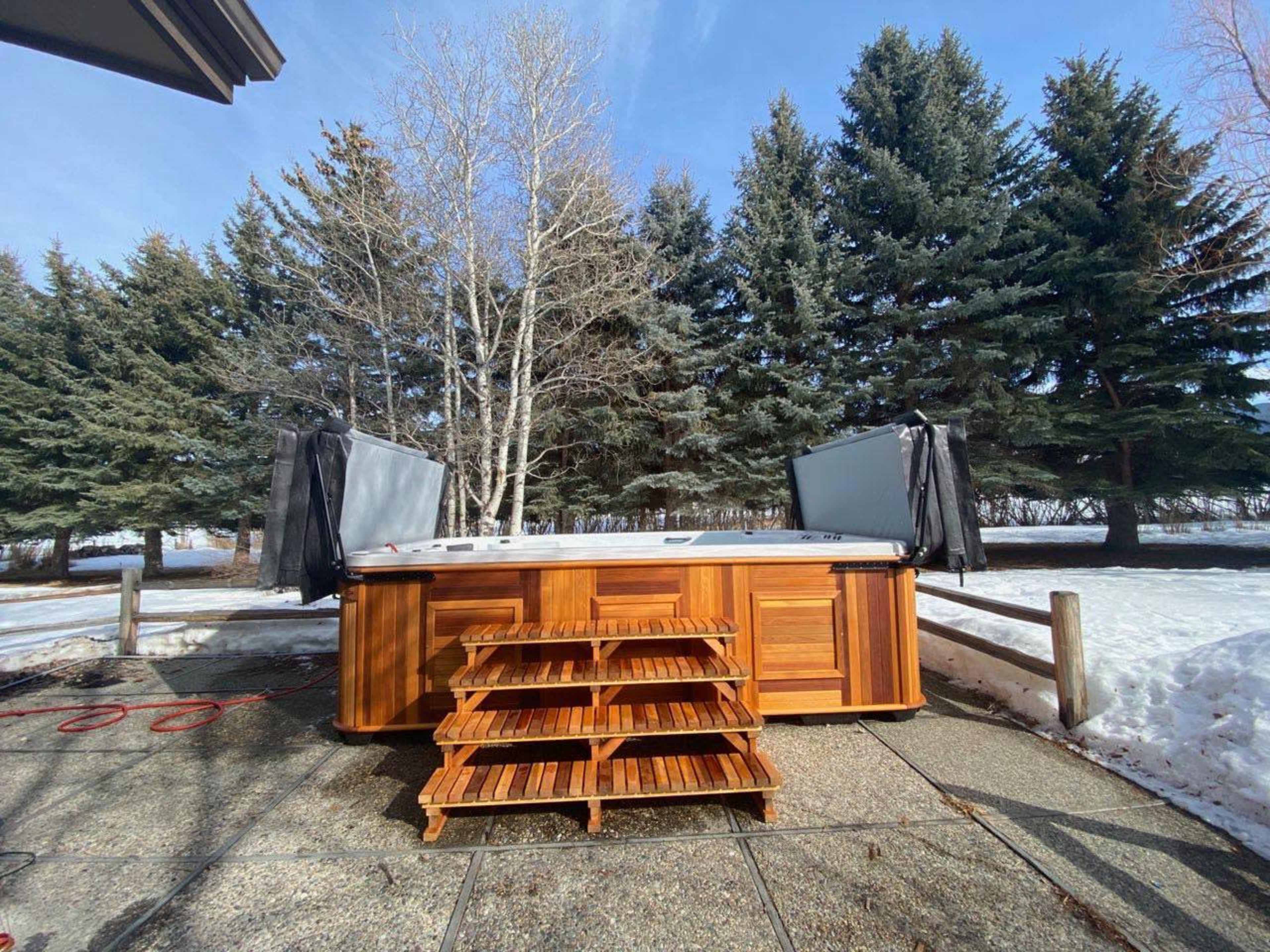 A wooden hot tub with steps is positioned on a stone patio surrounded by snowy ground and tall evergreen trees under a clear blue sky.