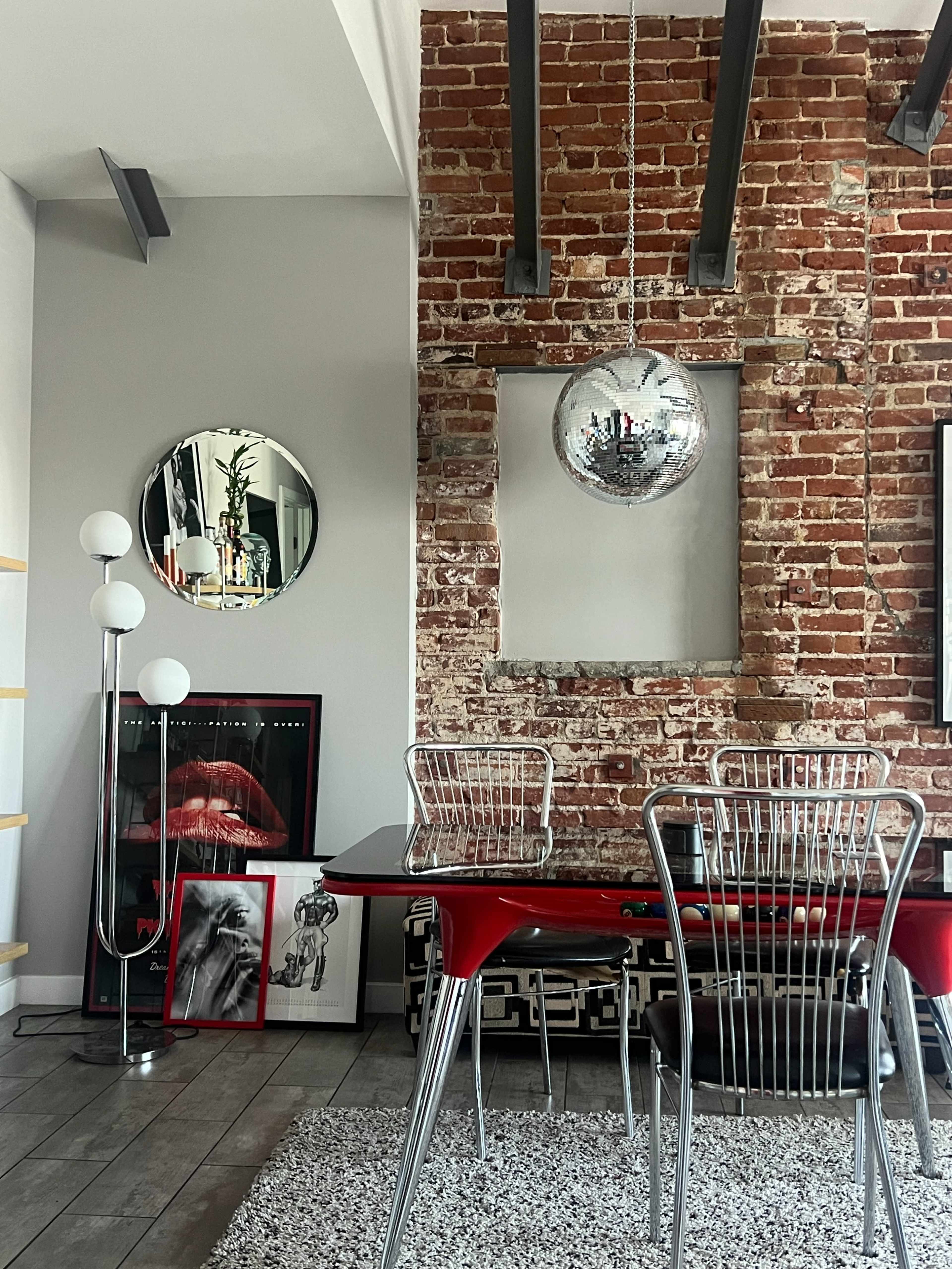 The image shows a modern dining area featuring a red table with metal chairs, a large disco ball hanging from the ceiling, and a textured brick wall adorned with mirrors and framed artwork.