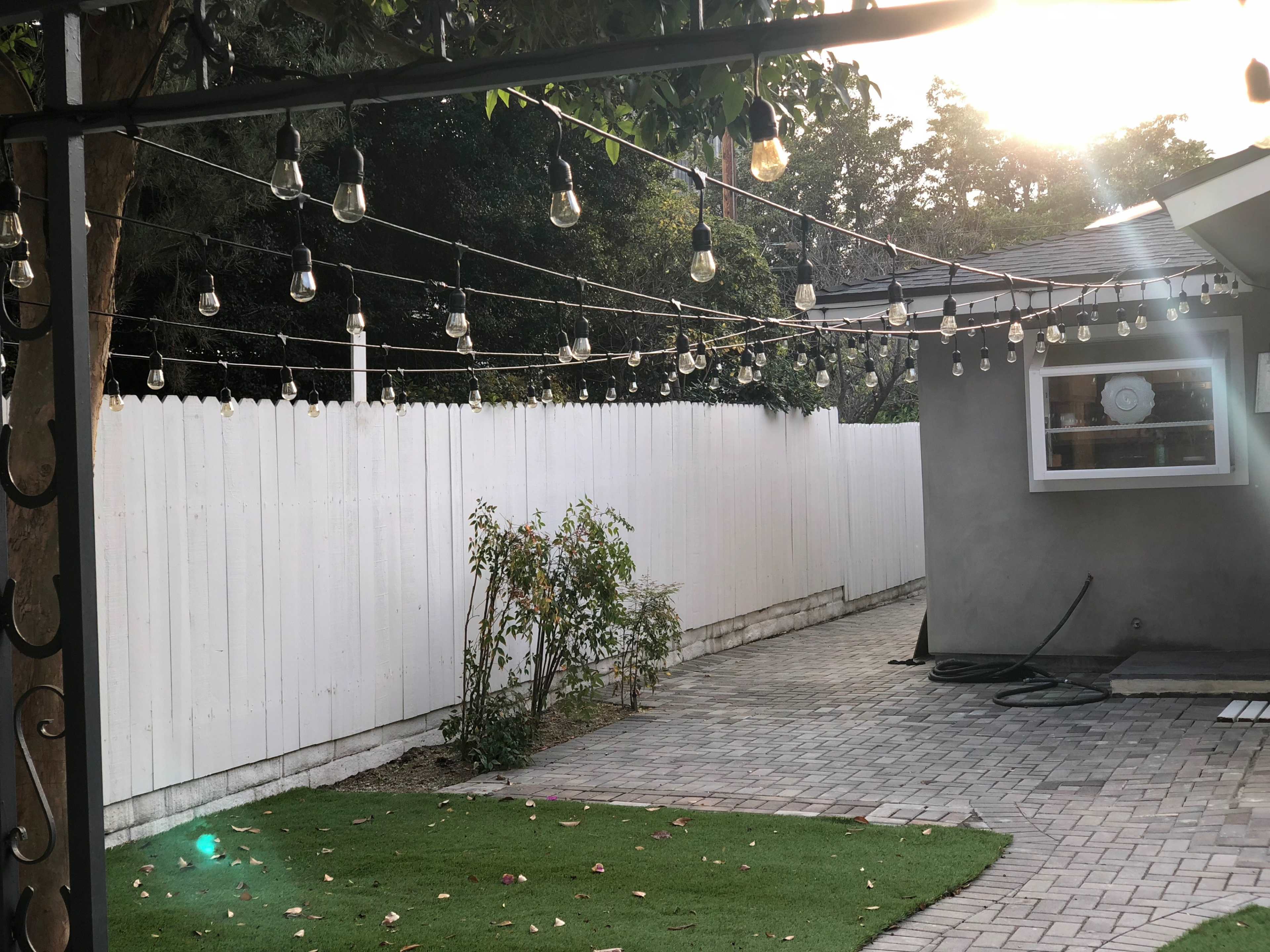 A backyard area with a paved path, a white fence, and string lights hanging overhead along with a house in the background.