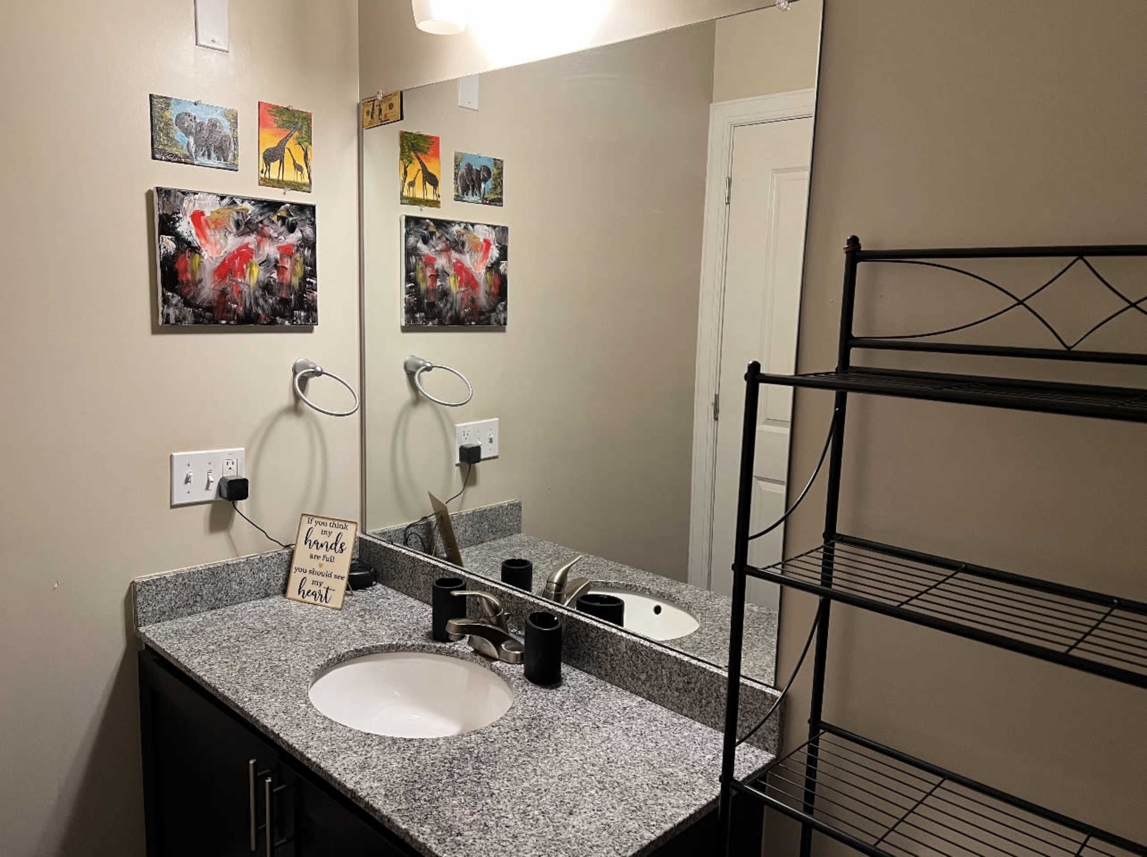 A bathroom features a granite countertop with a sink, a large mirror, two wall-mounted light fixtures, and artwork on the walls above.