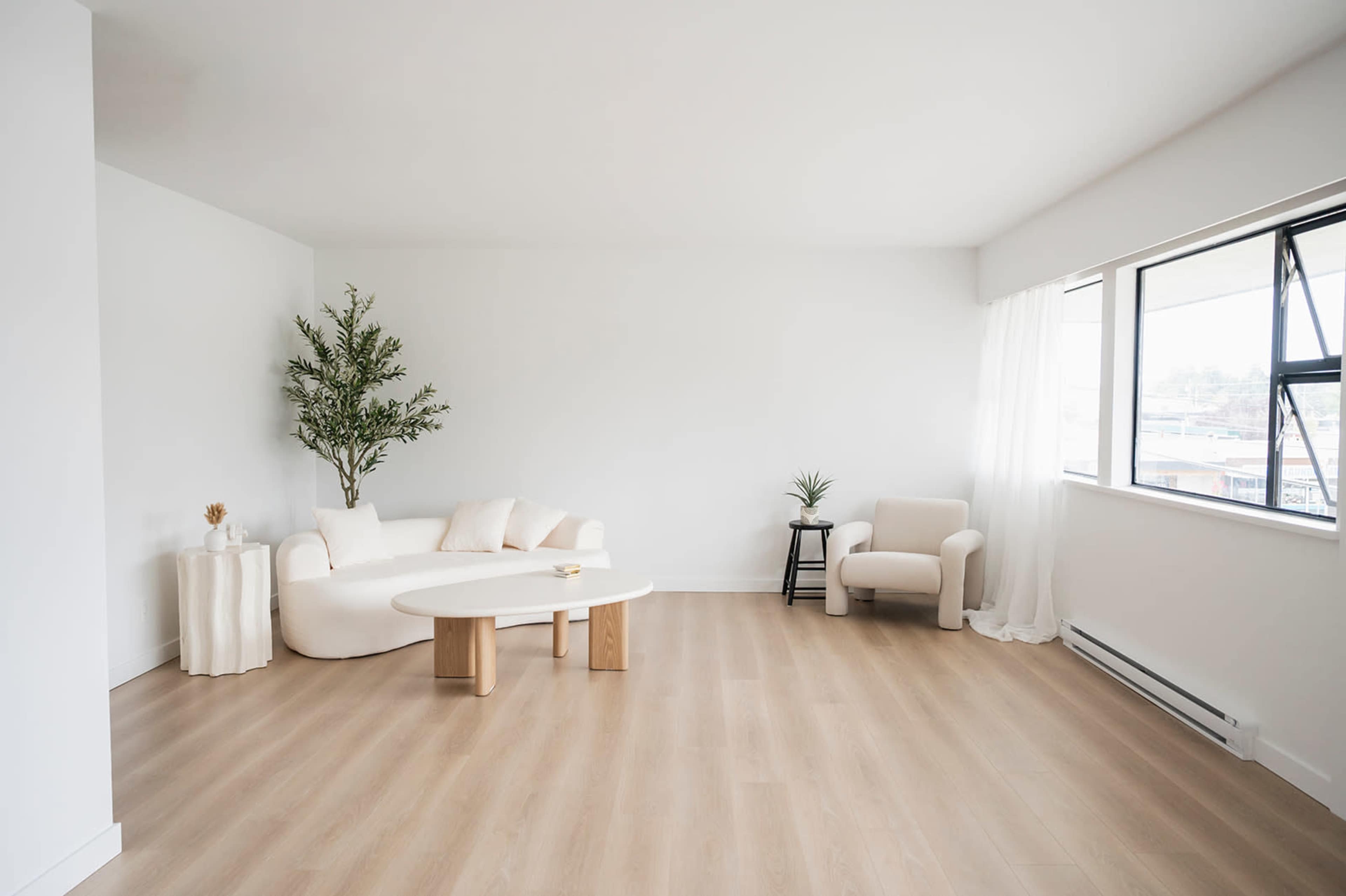 A brightly lit minimalist living room features a white sofa, a round white coffee table, and a beige armchair, with a small indoor tree and decorative plants.
