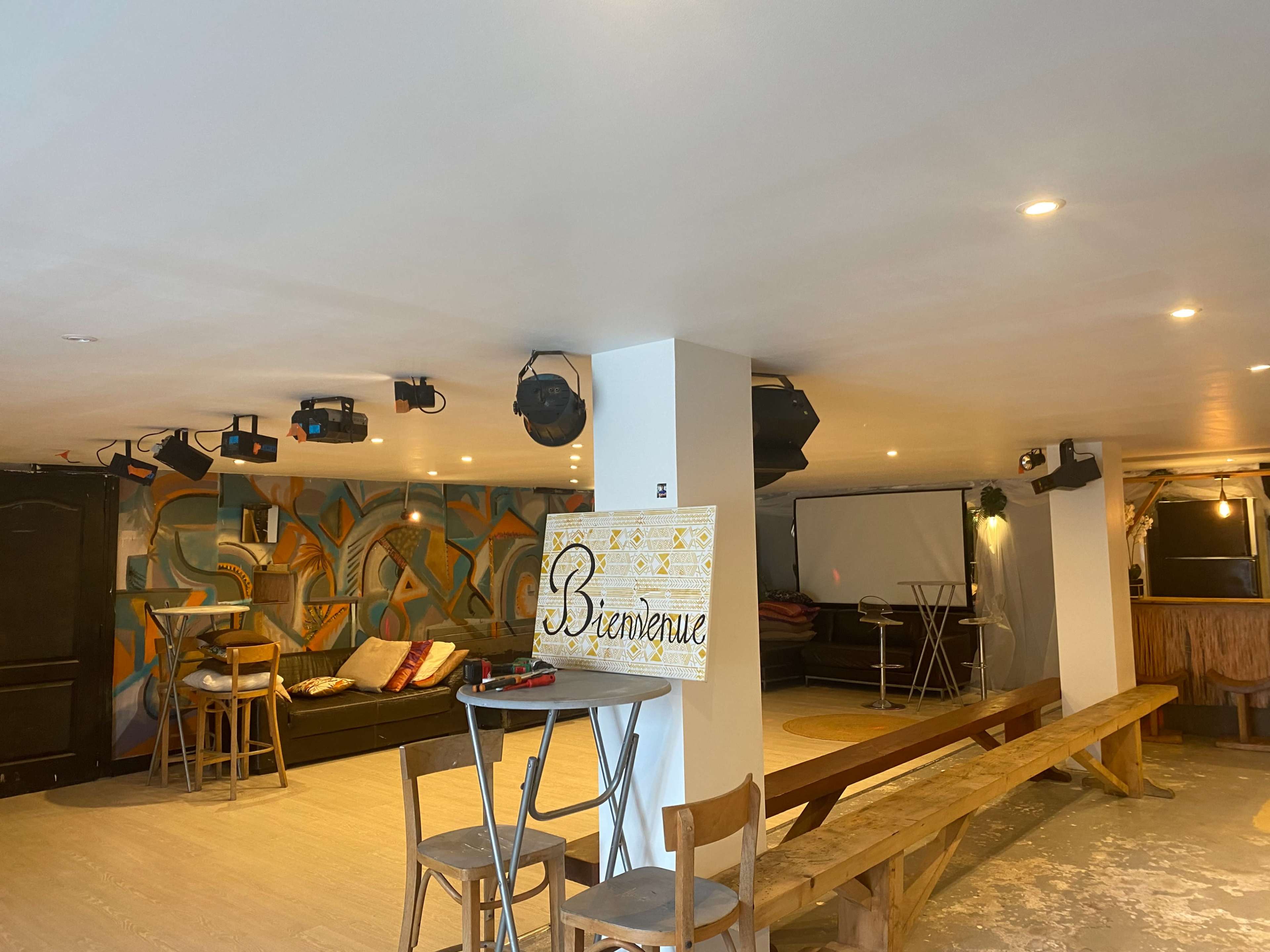 A spacious, dimly lit lounge area features benches and tables with artistic wall decor and a projector screen in the background.