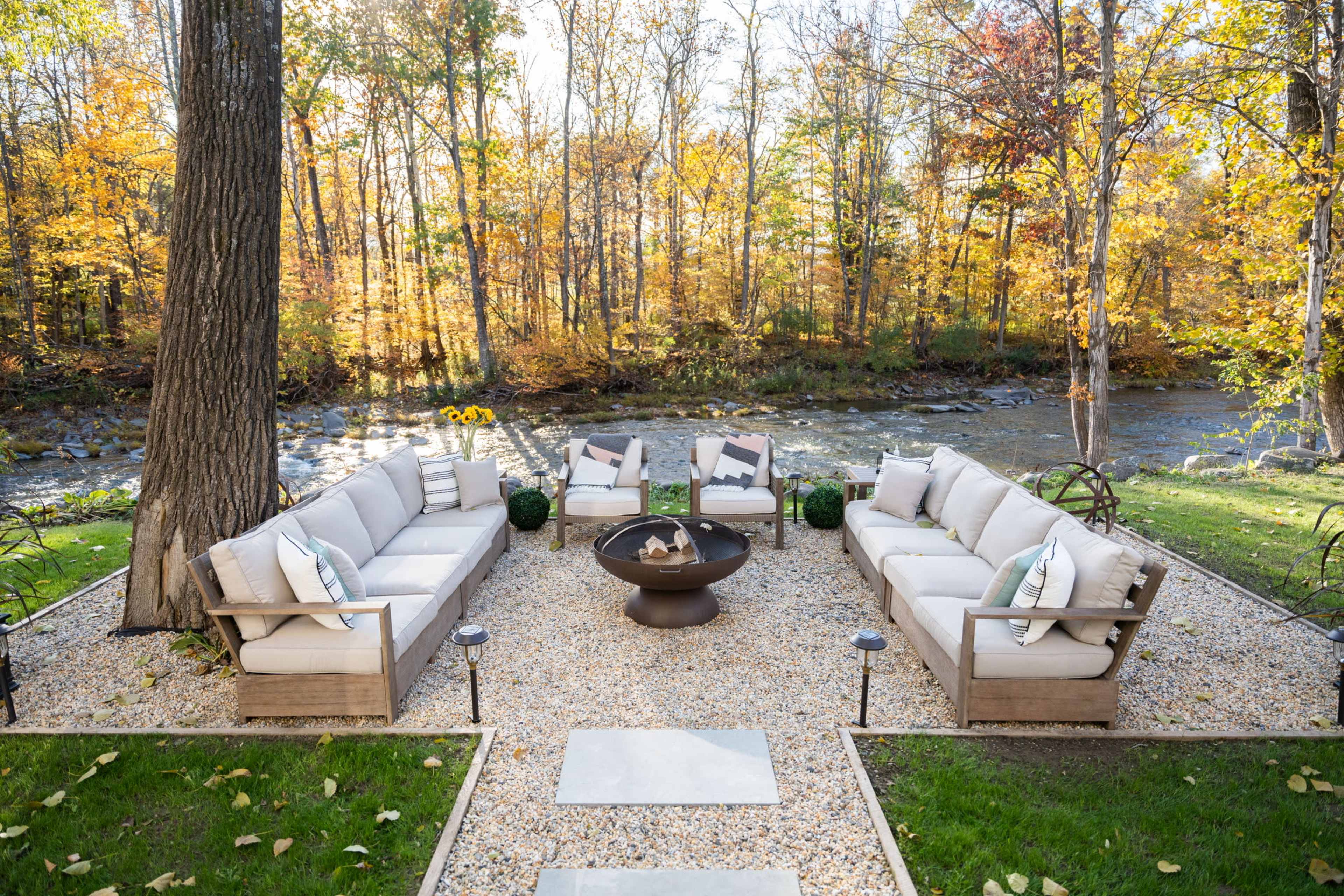 A cozy outdoor seating area is arranged around a fire pit, with a backdrop of trees displaying autumn foliage near a flowing river.
