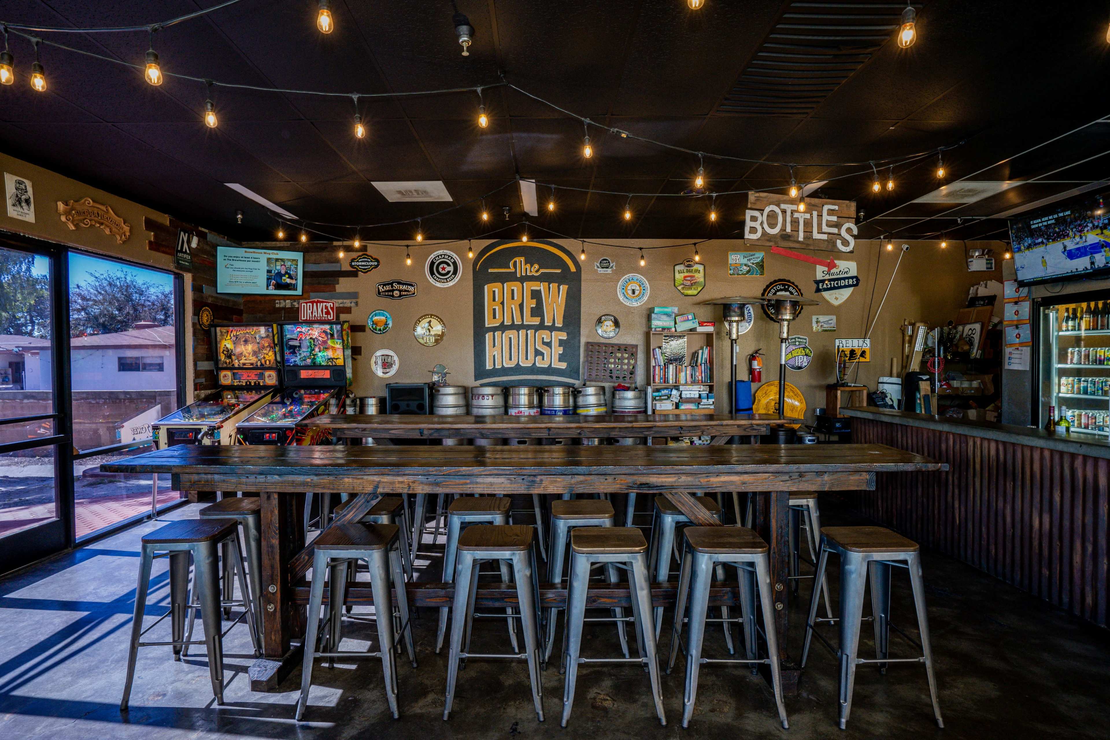 The interior of The Brew House features a long wooden bar with metal stools, illuminated by string lights and adorned with vintage signs and a variety of beverages.