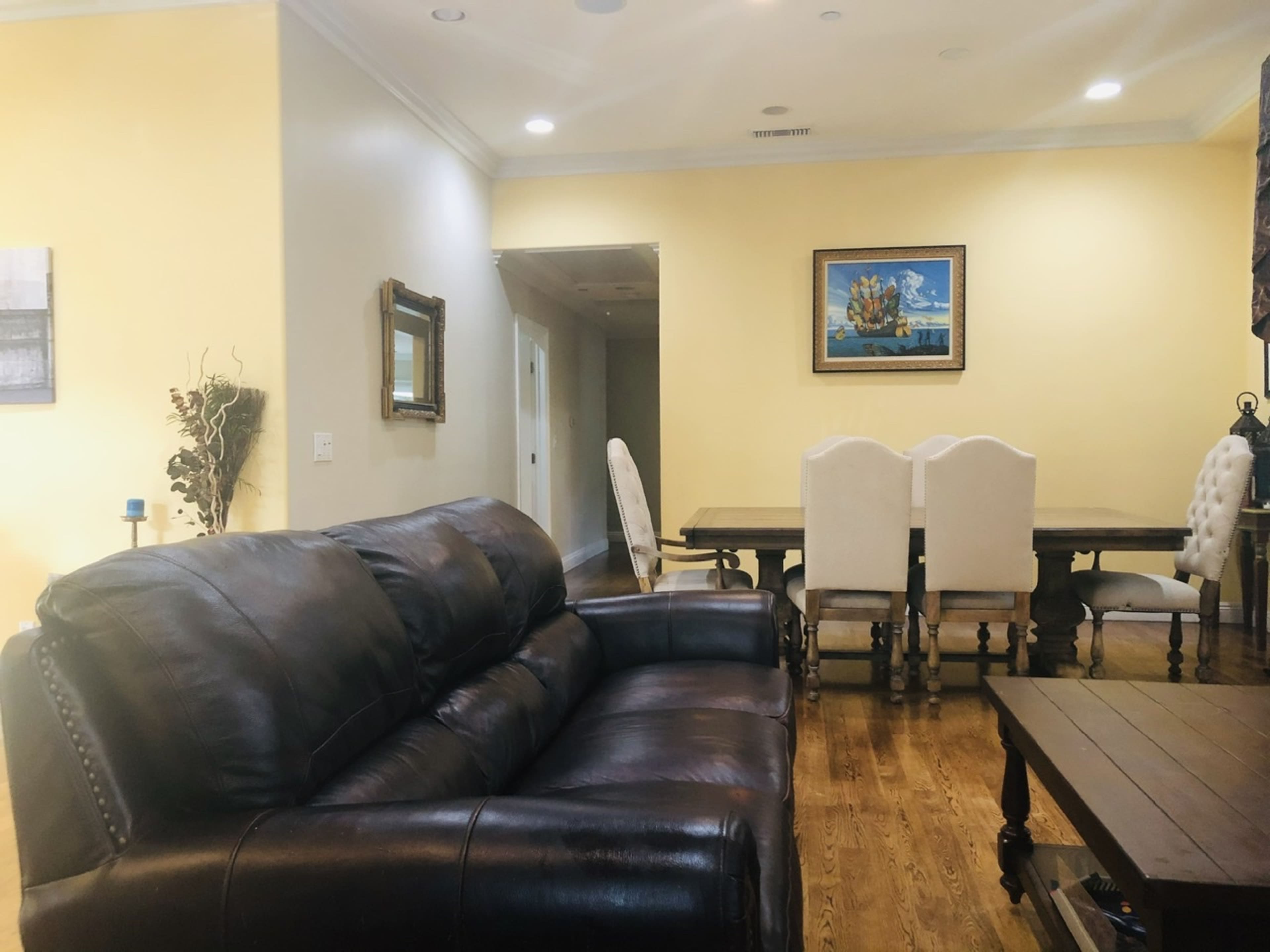 A brown leather couch facing a dining area with a wooden table and white chairs in a yellow-walled room.