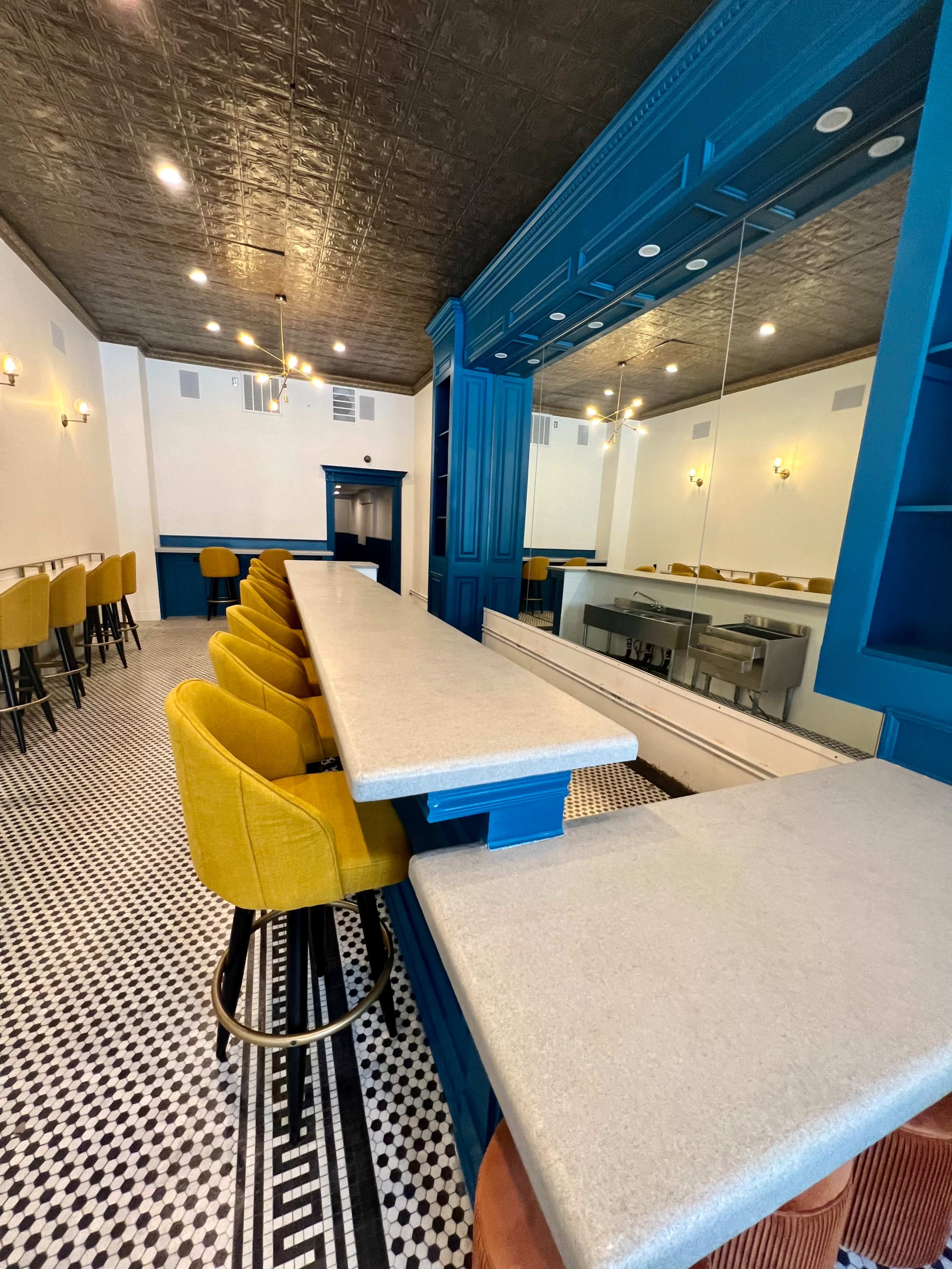 A modern bar area featuring yellow upholstered stools along a light gray countertop, with a blue paneled wall and a patterned black-and-white floor.