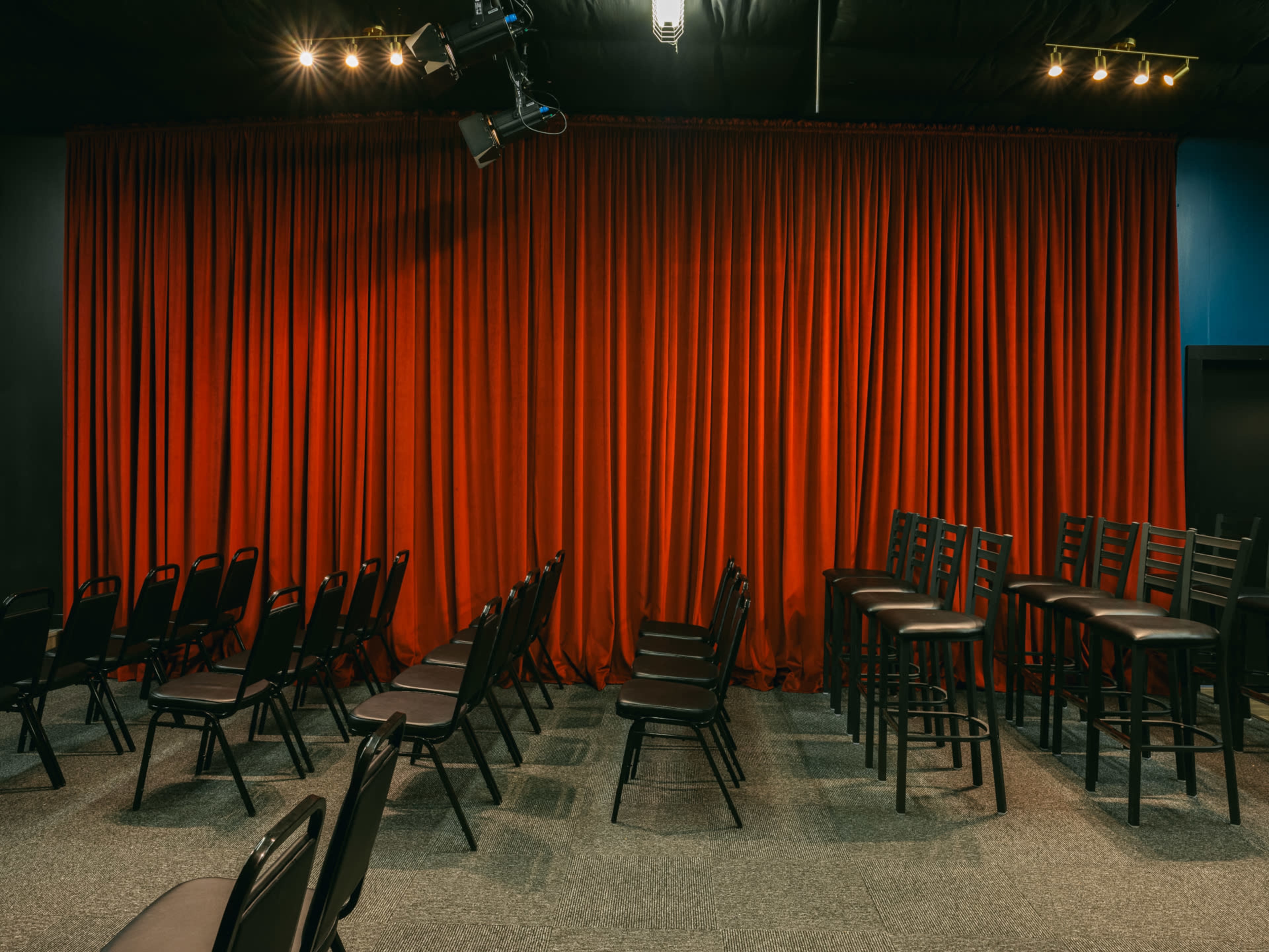 A small performance space with rows of black chairs facing a red curtain, alongside stacked black bar stools.