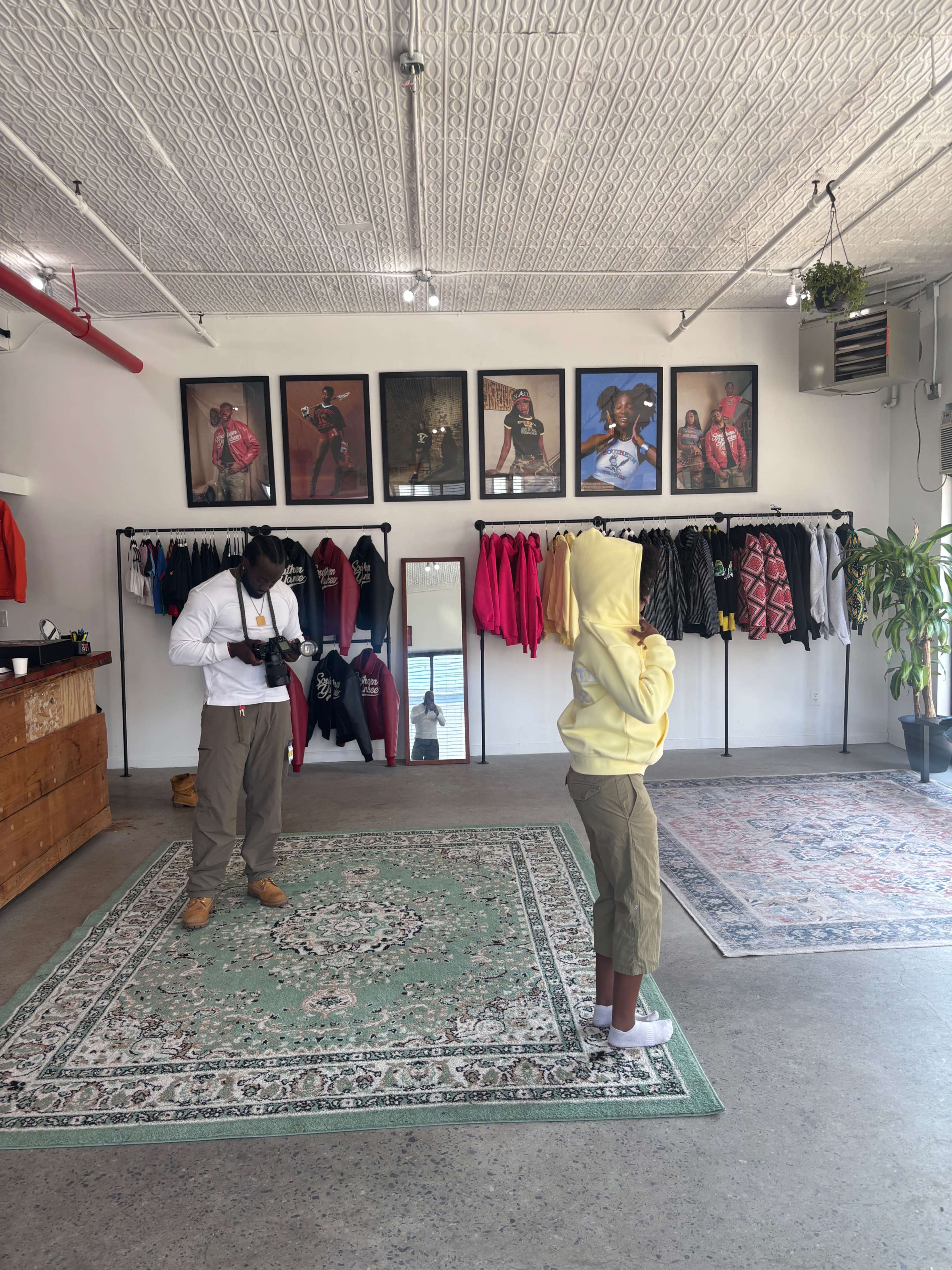 A photographer captures an image of a child wearing a yellow hoodie, standing in a clothing store filled with various garments and framed artworks on the walls.