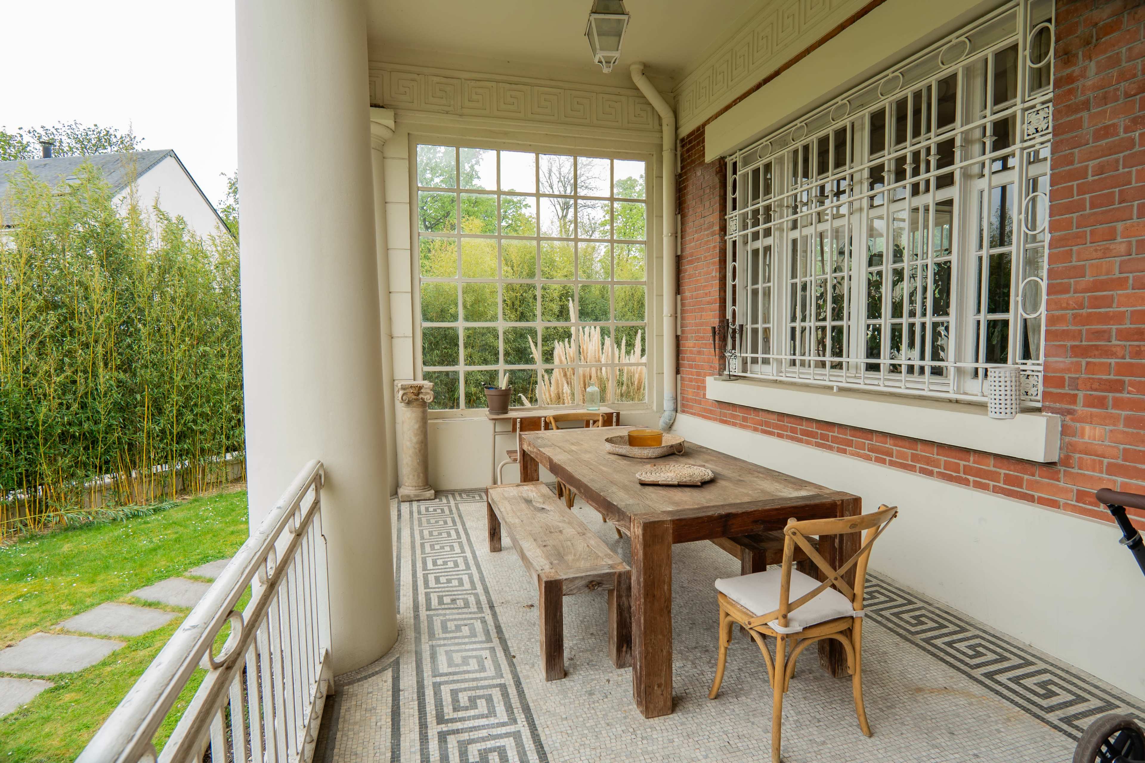 The image shows a covered patio with a wooden table, chairs, and a decorative wall, framed by large windows and surrounded by greenery.
