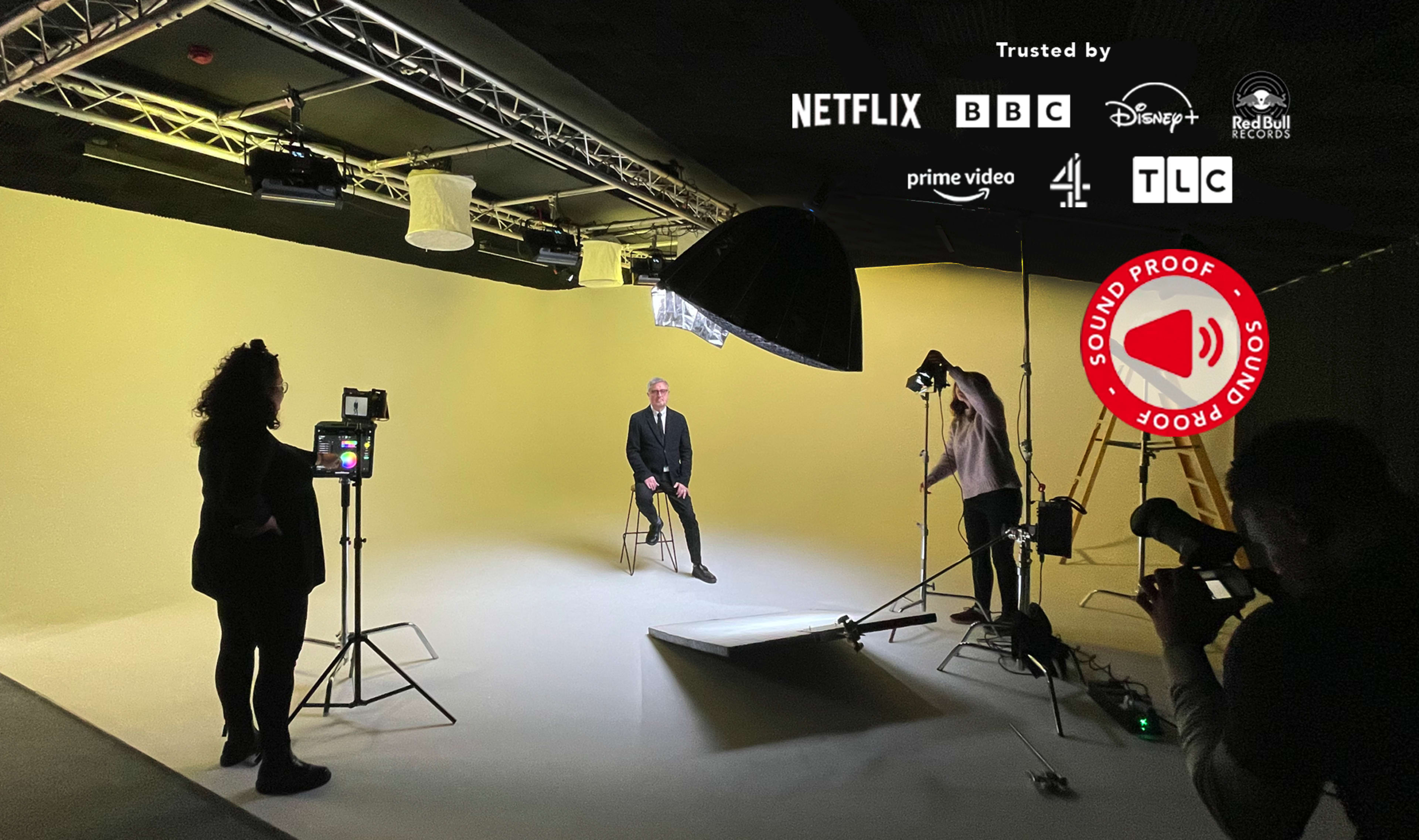 A studio setup features a man in a suit sitting on a chair in front of a yellow backdrop, with various lighting equipment and cameras positioned around him.