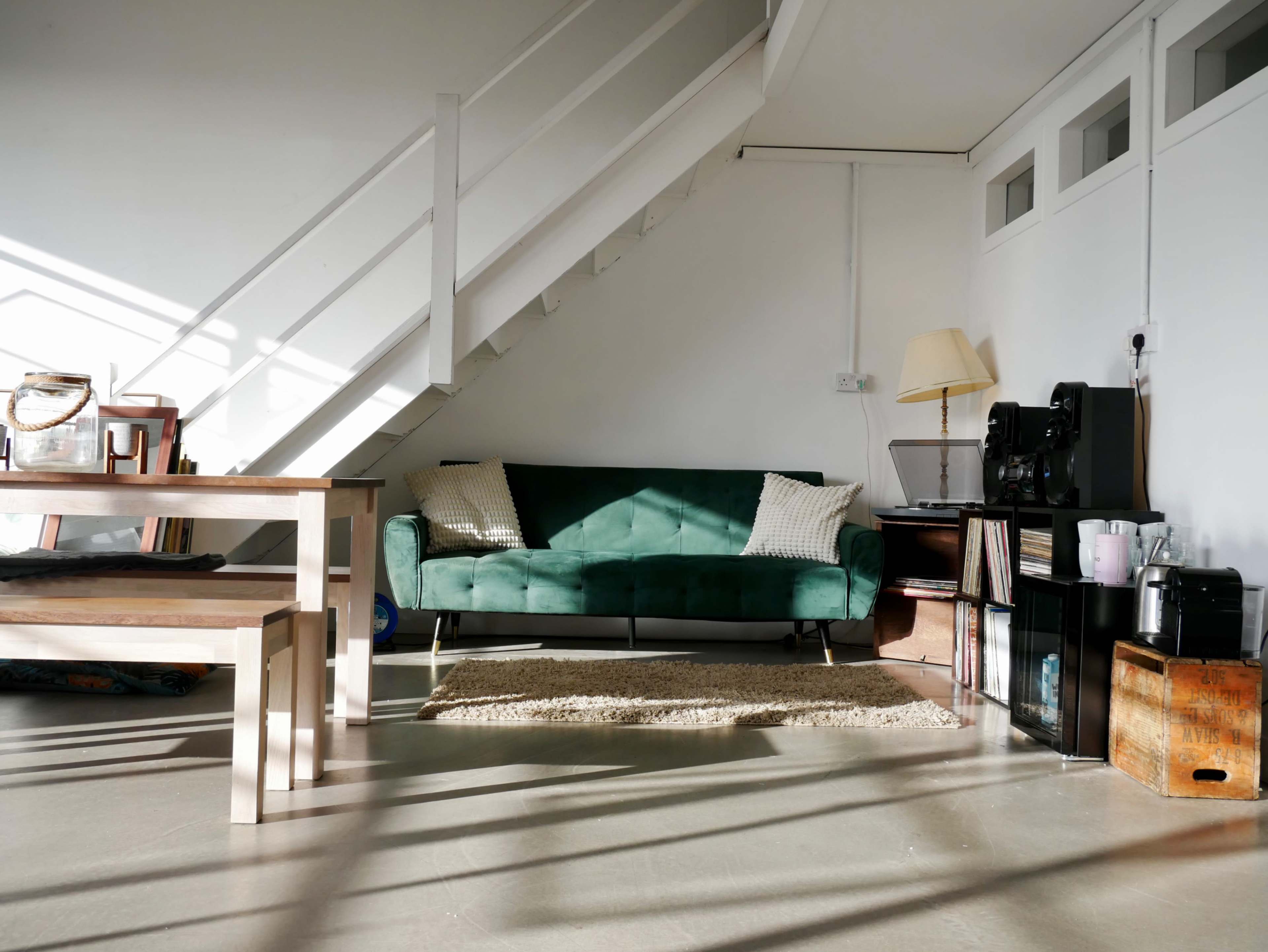 The image shows a modern living area with a green sofa, a wooden table, a staircase, and various furnishings along the walls.