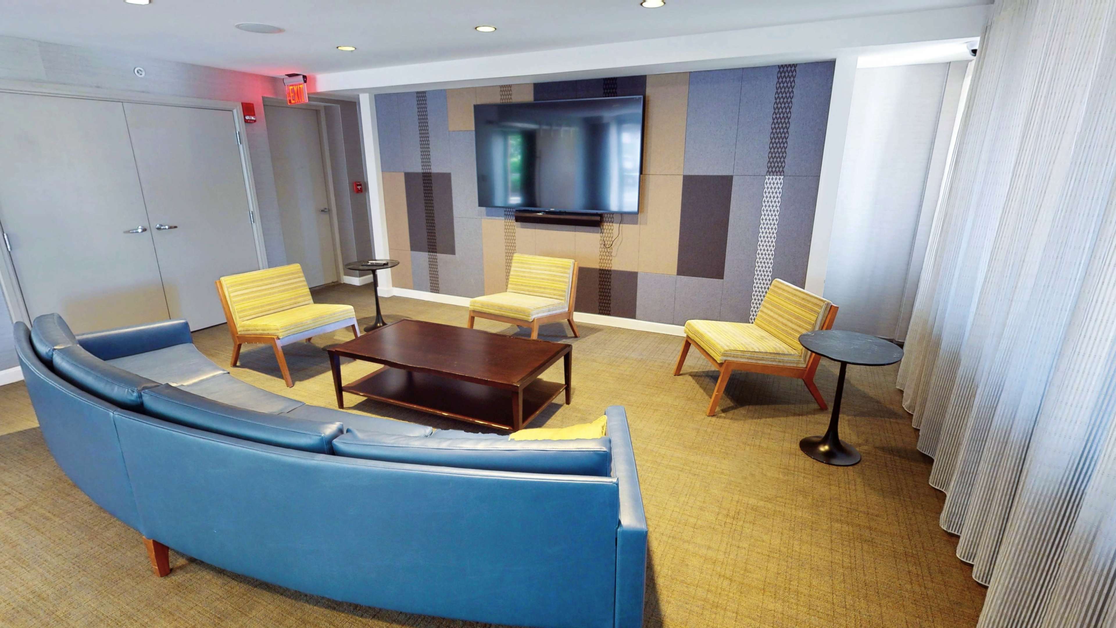 The image shows a modern lounge area featuring a curved blue sofa, four yellow chairs, a coffee table, and a wall-mounted television.
