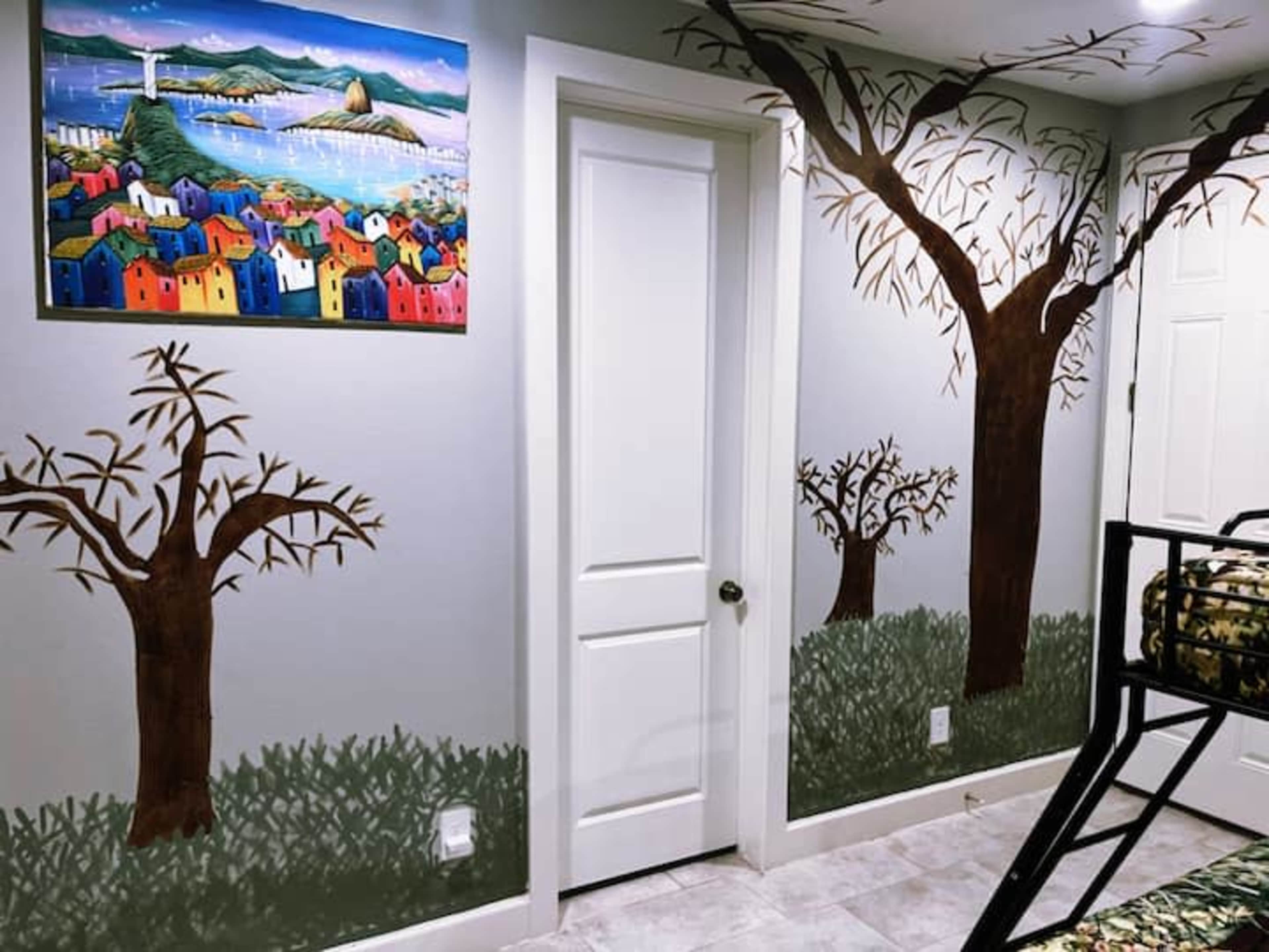 A room with mural-painted trees and colorful houses, with a door and a large painting on the wall.