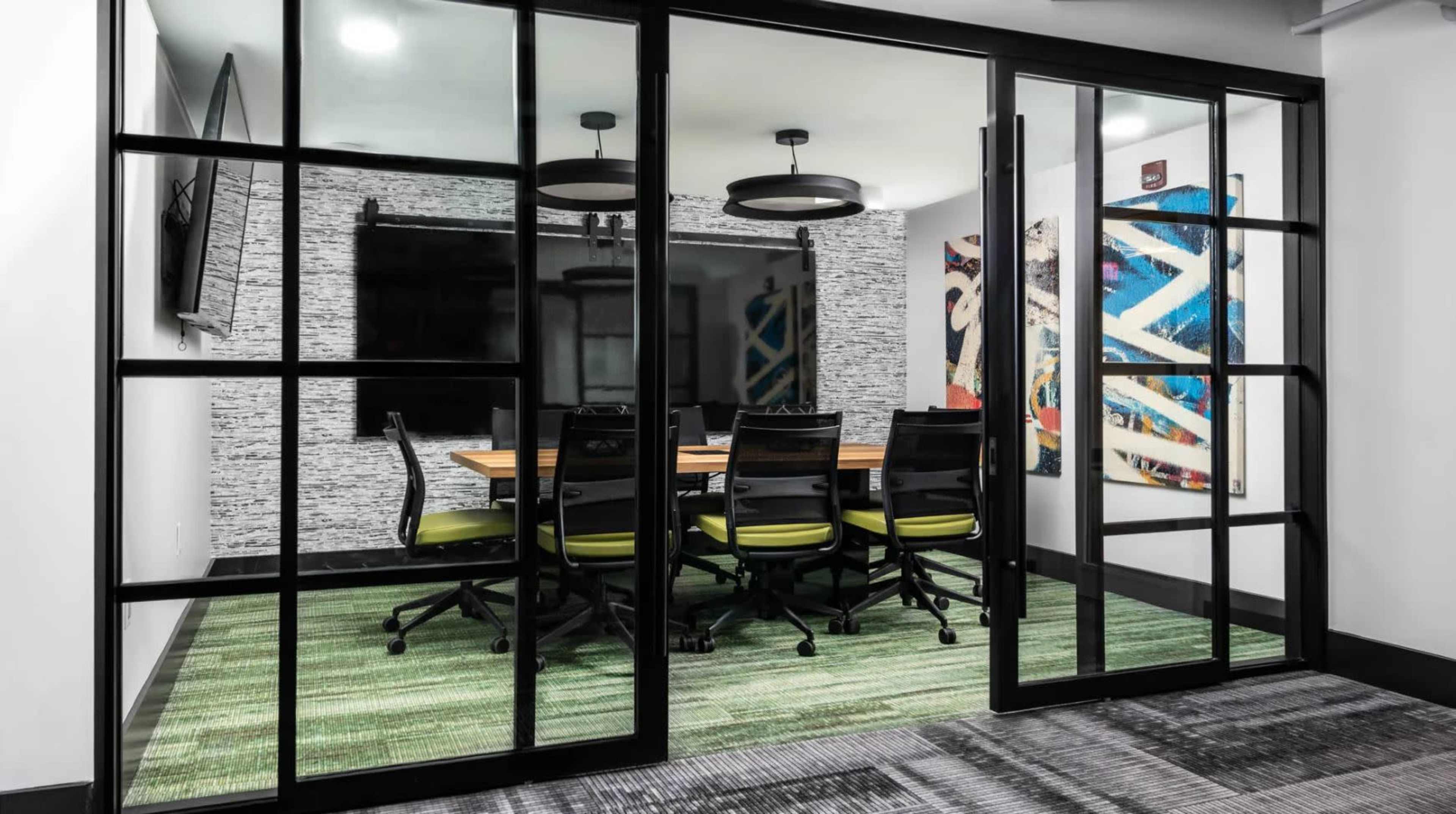 A glass-walled conference room features a wooden table surrounded by six black chairs with green accents, set against a backdrop of abstract artwork and a television screen.