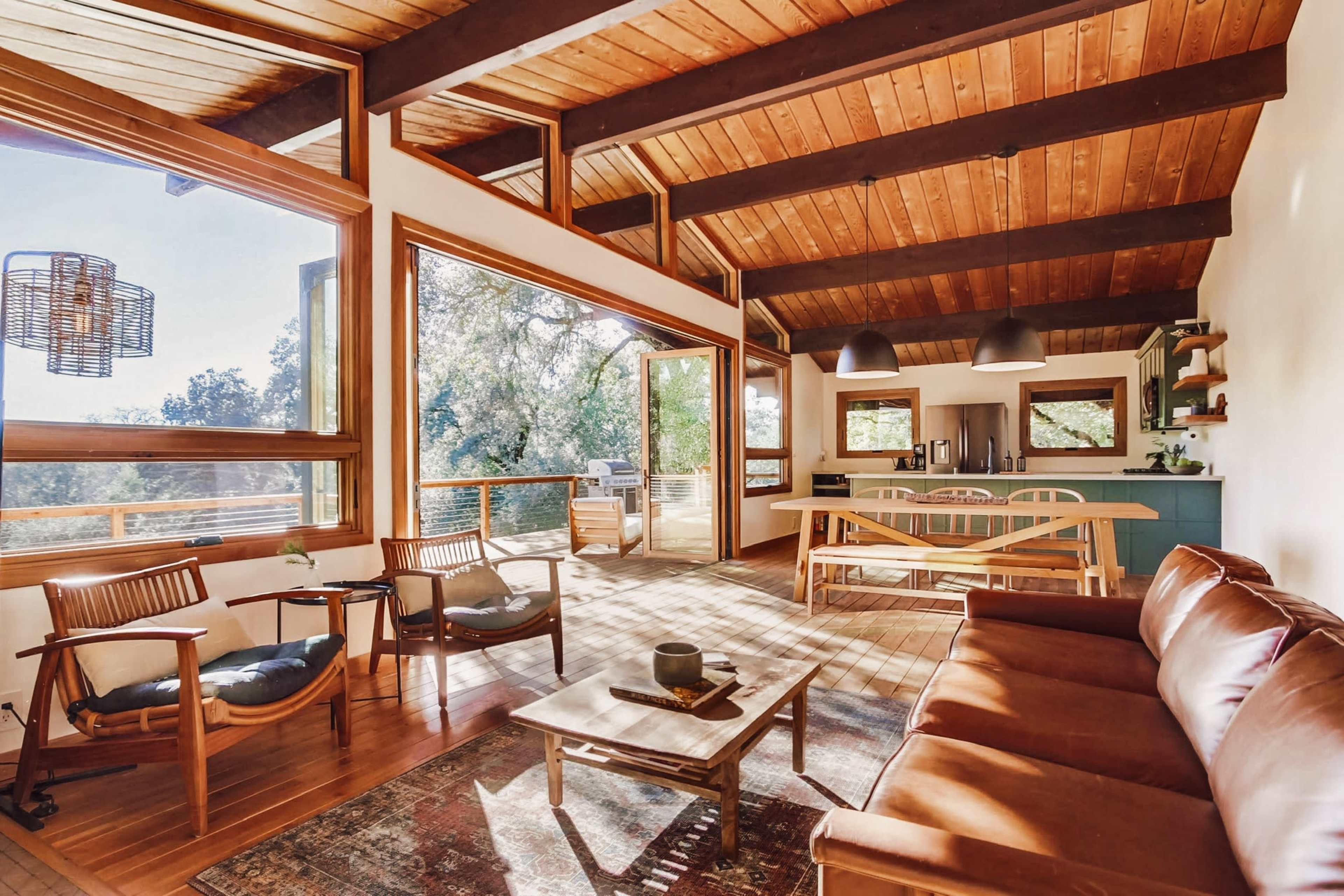 The scene shows a spacious, modern living area with large windows, wooden beams on the ceiling, a leather couch, and a dining area leading to an outdoor deck.