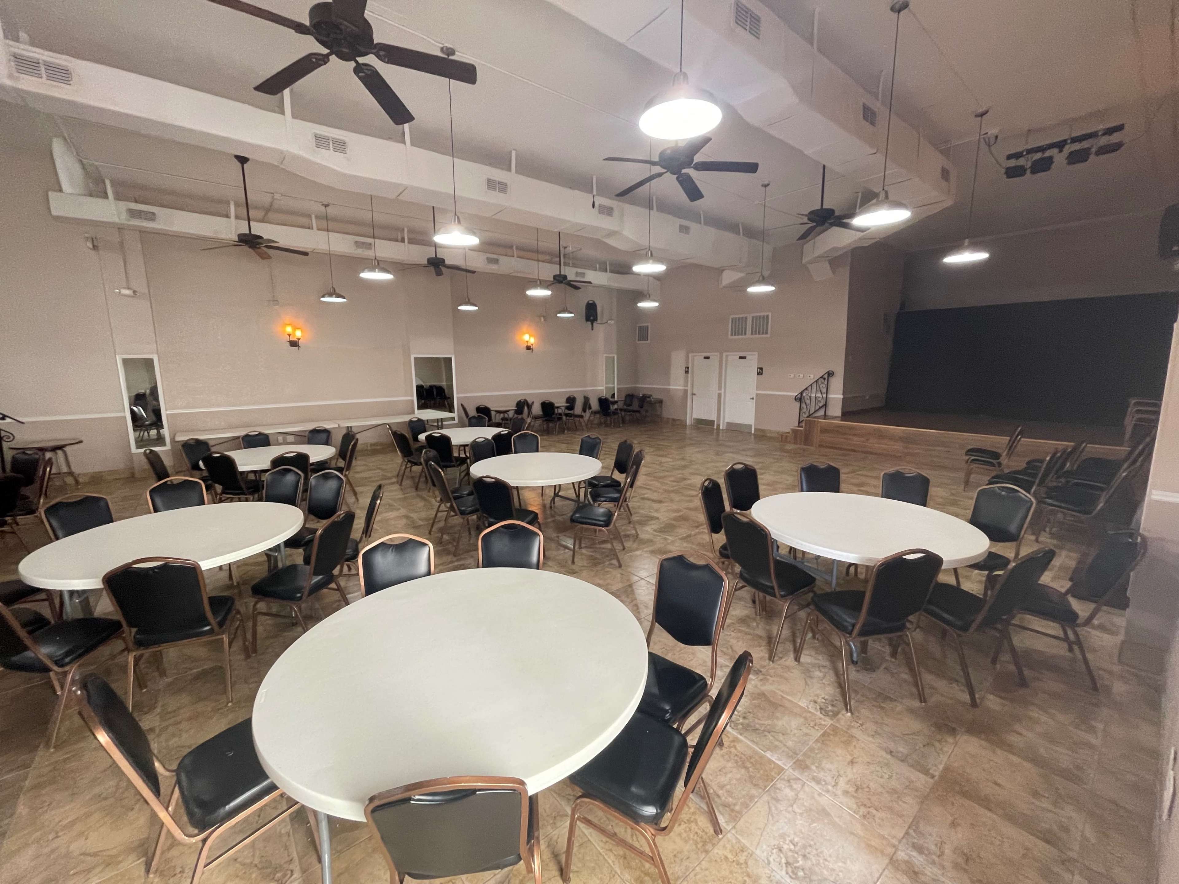 The image shows an empty indoor event space with multiple round tables and chairs arranged around the room, and a stage at the far end.
