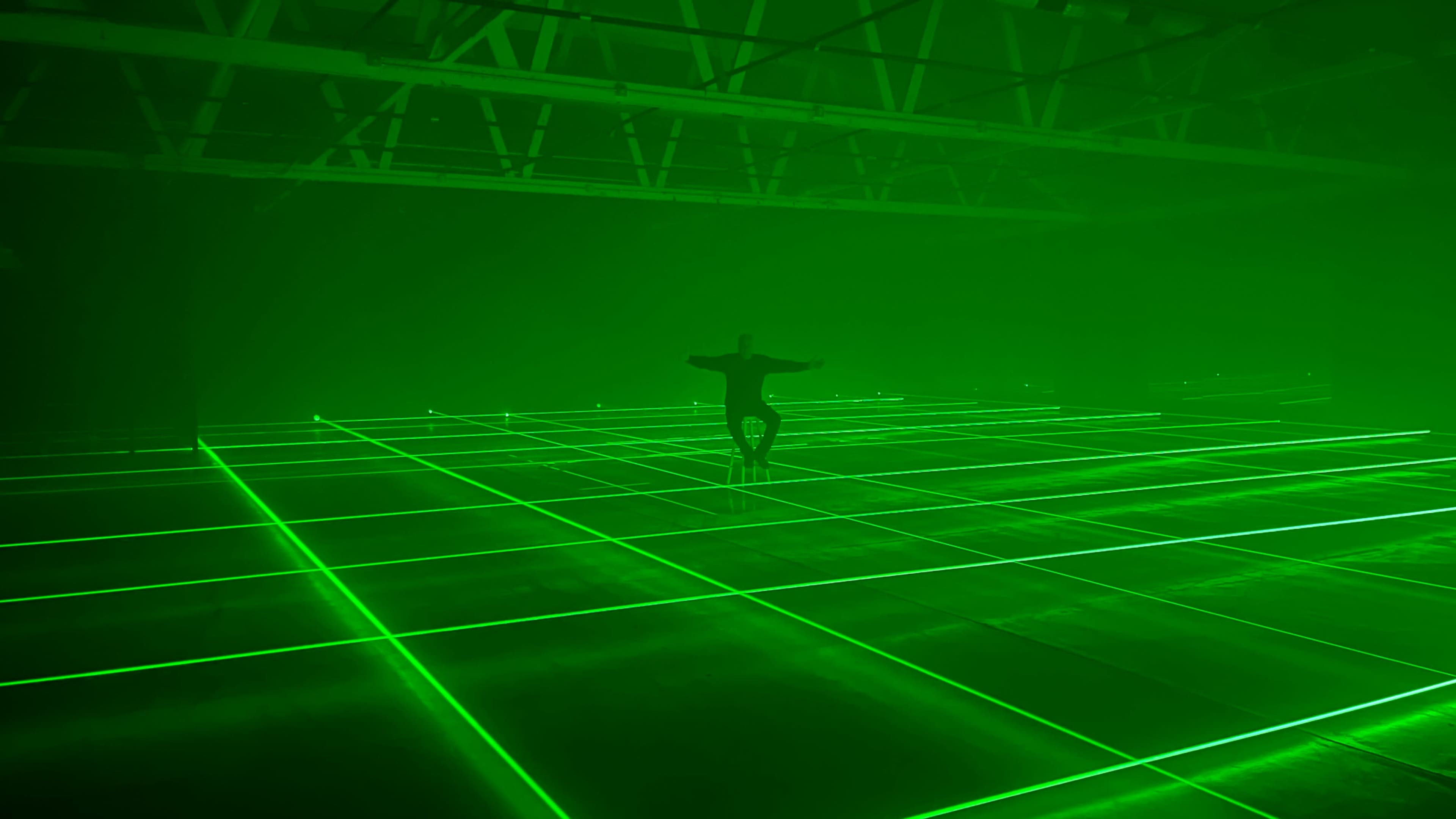 A person stands with arms outstretched on a grid of green laser lines in a dark, expansive space.