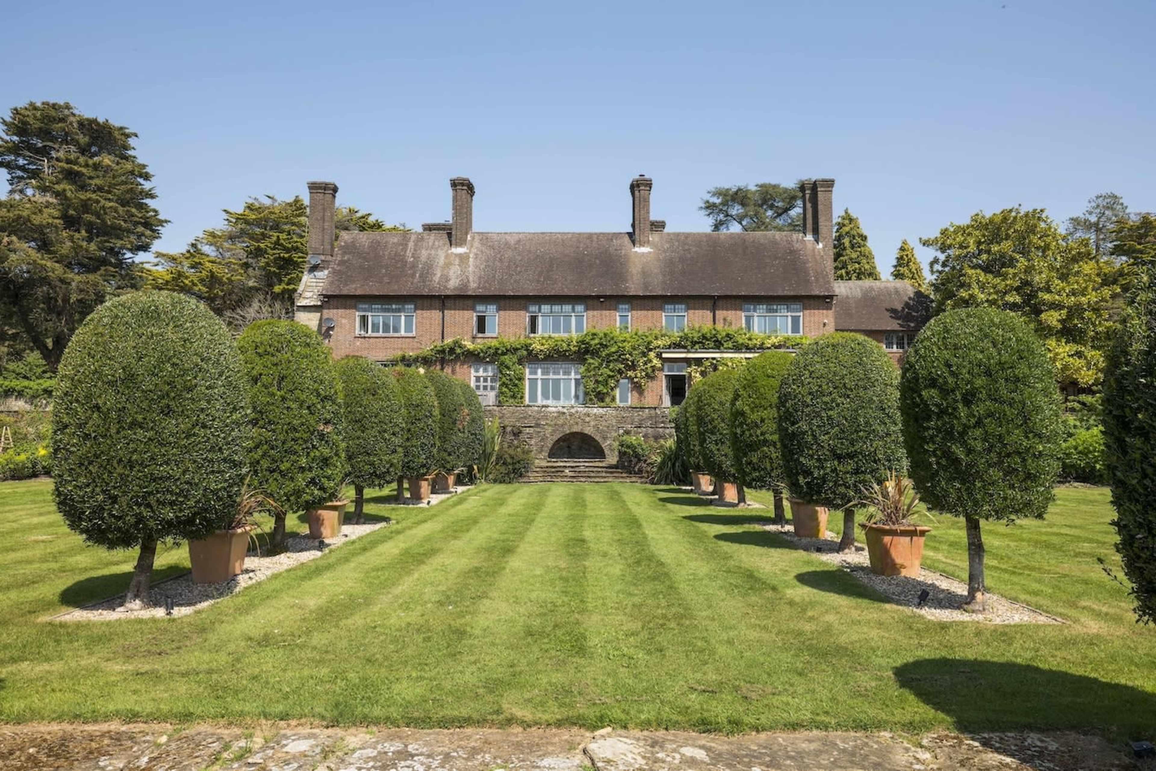 A large brick house is set behind a manicured lawn flanked by neatly trimmed topiary trees in pots.