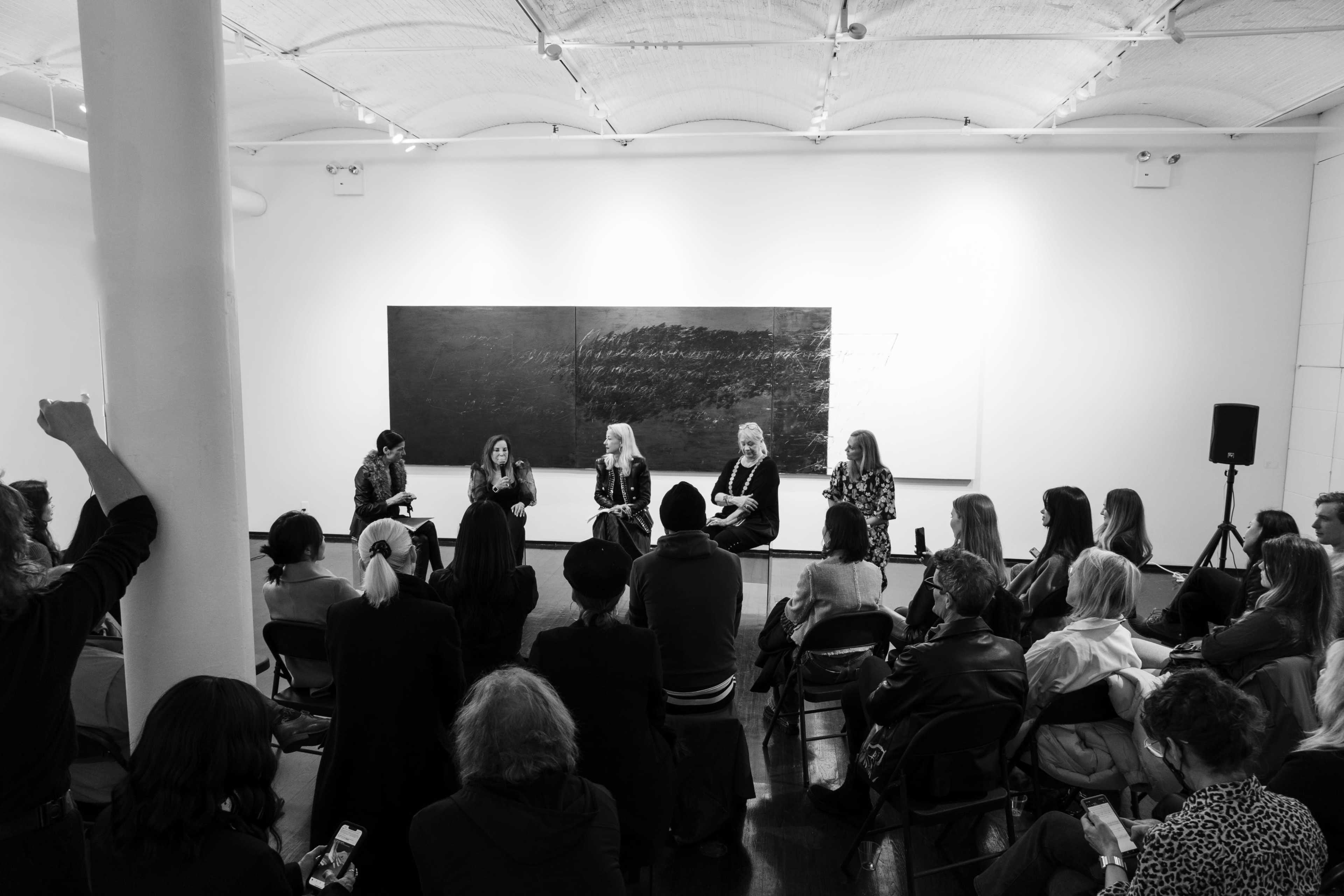 A panel discussion takes place in a gallery with four speakers seated in front of an audience.