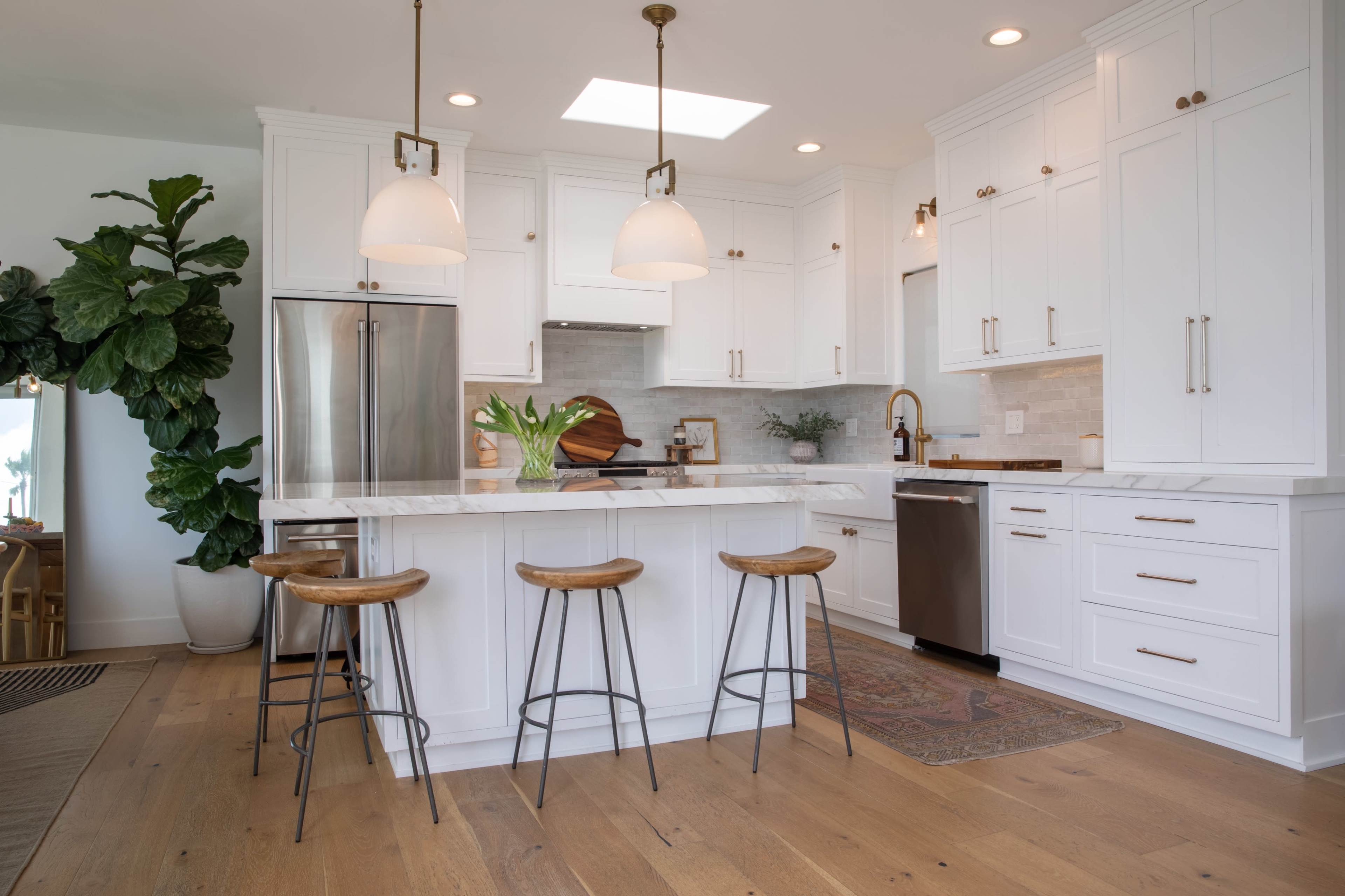 A modern kitchen features a white cabinetry setup with a central island and three wooden stools, complemented by pendant lighting and large windows.