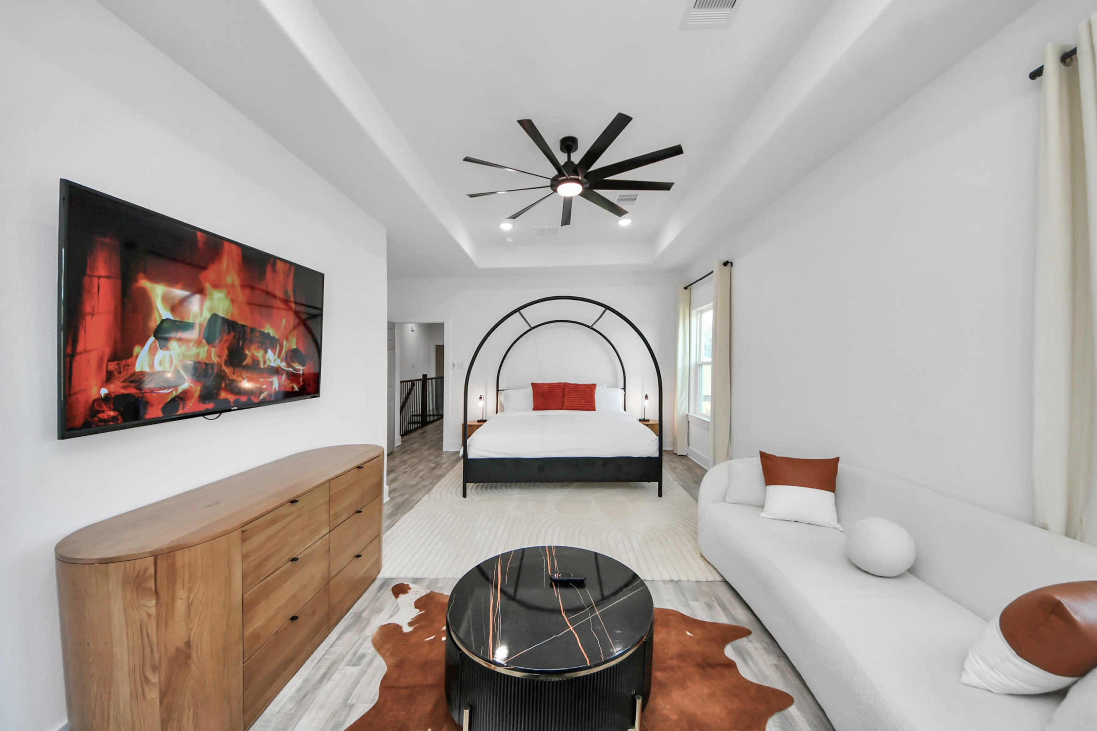 The image shows a modern bedroom featuring a large bed with a curved headboard, a cozy seating area with a curved sofa and round coffee table, and a wall-mounted television displaying a fireplace.