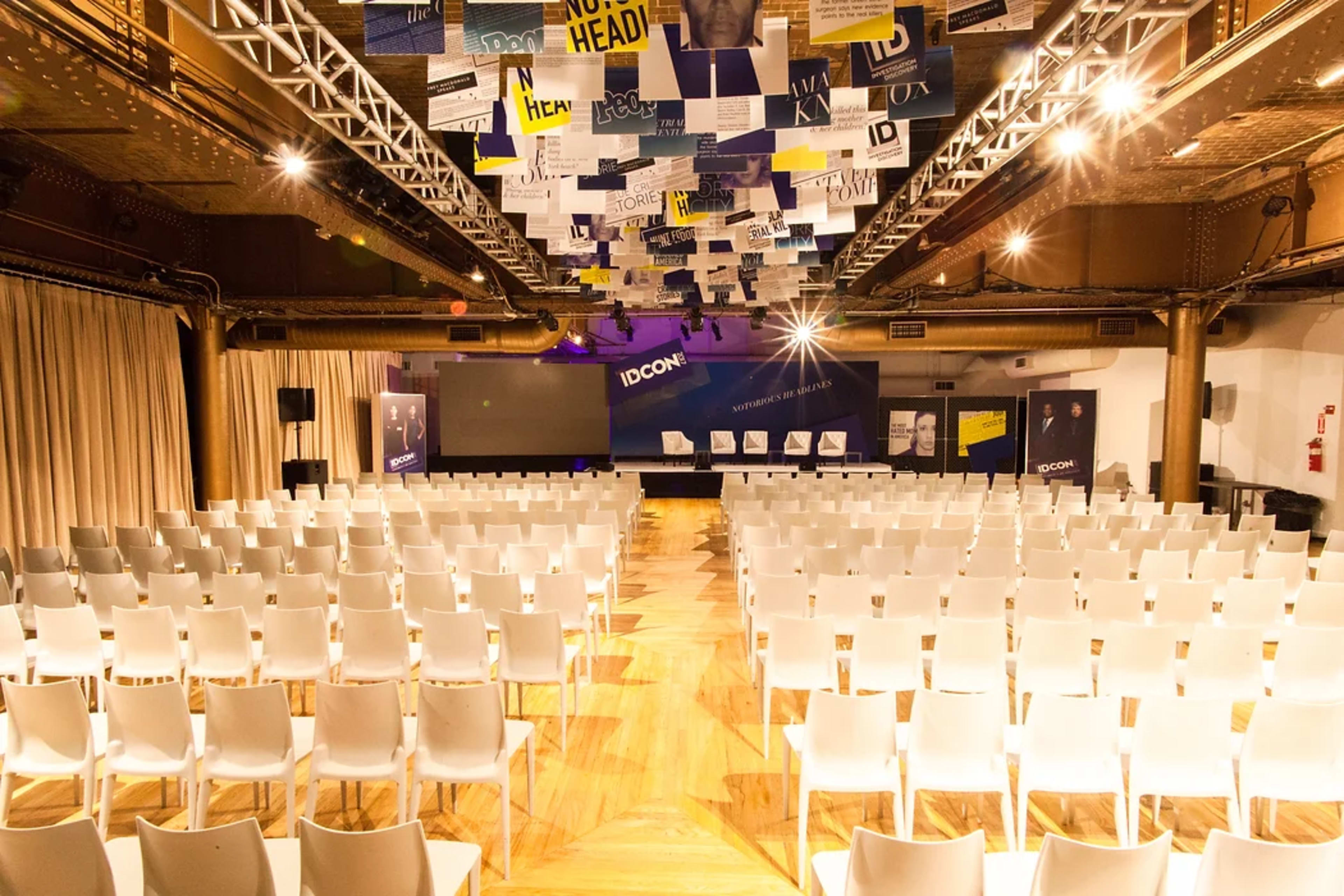 A conference hall is set up with rows of white chairs facing a stage adorned with banners and lights.