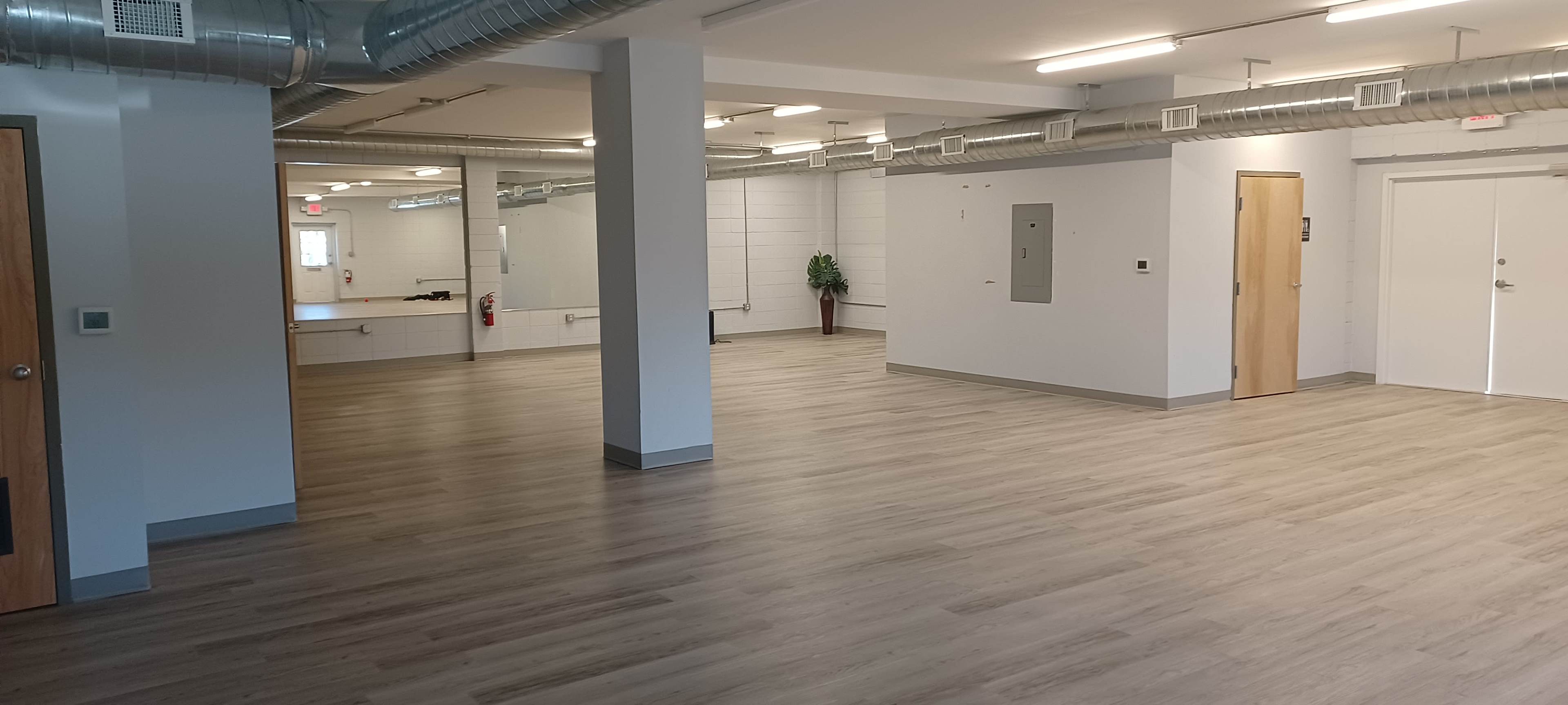 The image shows an empty interior space with wooden flooring, large windows, and a few doors leading to other areas.