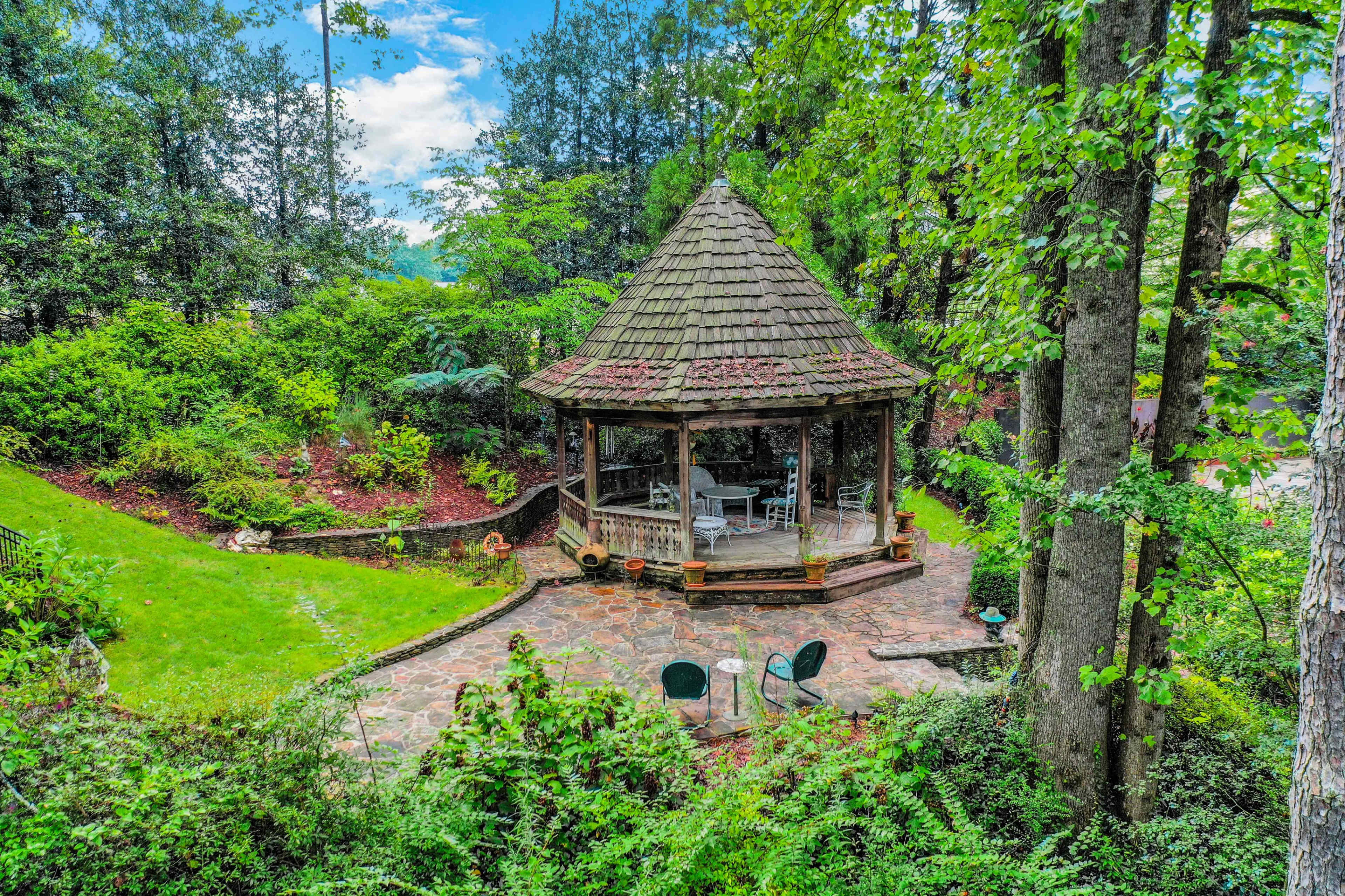 A wooden gazebo with a shingled roof is surrounded by greenery and stone pathways in a landscaped garden.