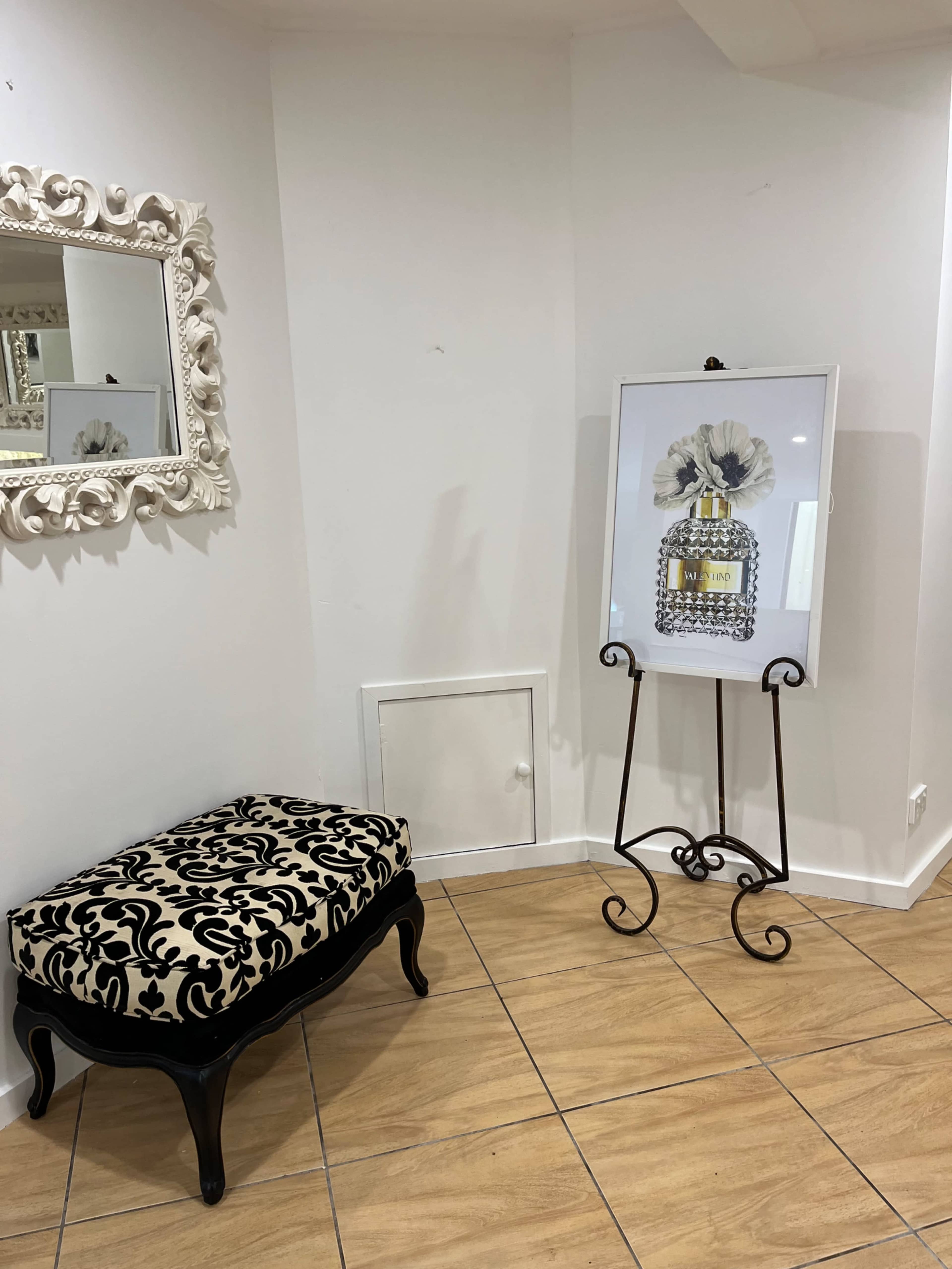 The image shows a corner of a room featuring a decorative bench with a patterned cushion, a framed artwork on an easel, and a wall mirror.