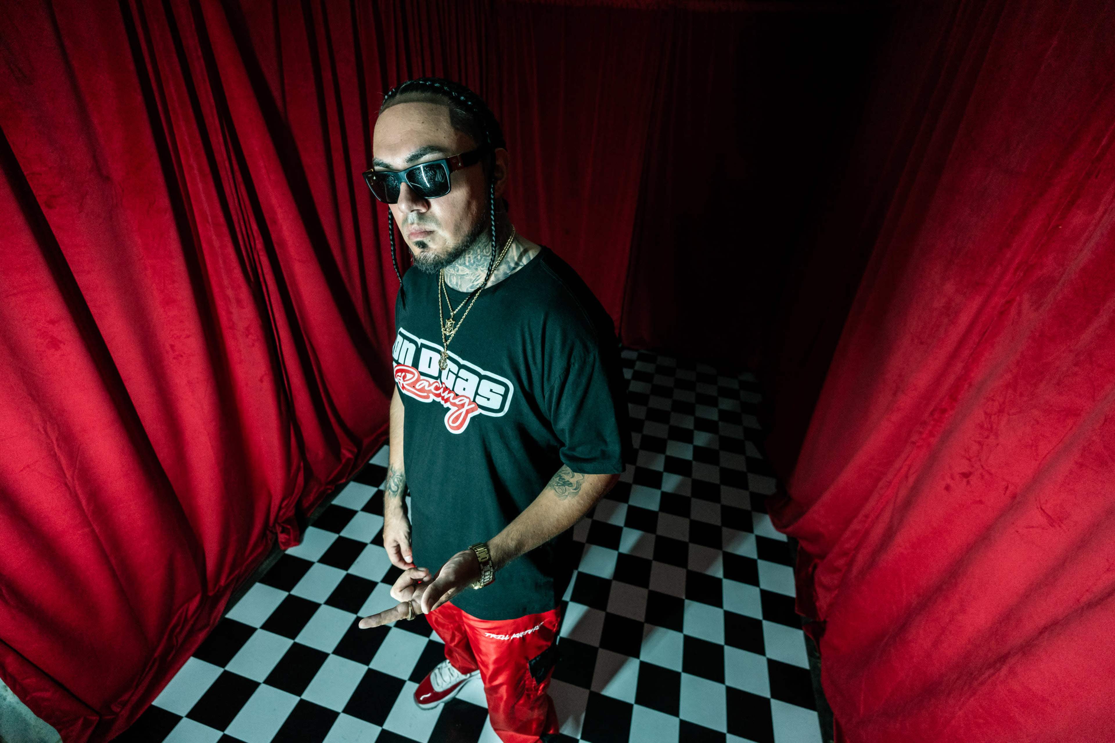 A man wearing sunglasses and a black t-shirt stands in a room with red curtains and a checkered floor.
