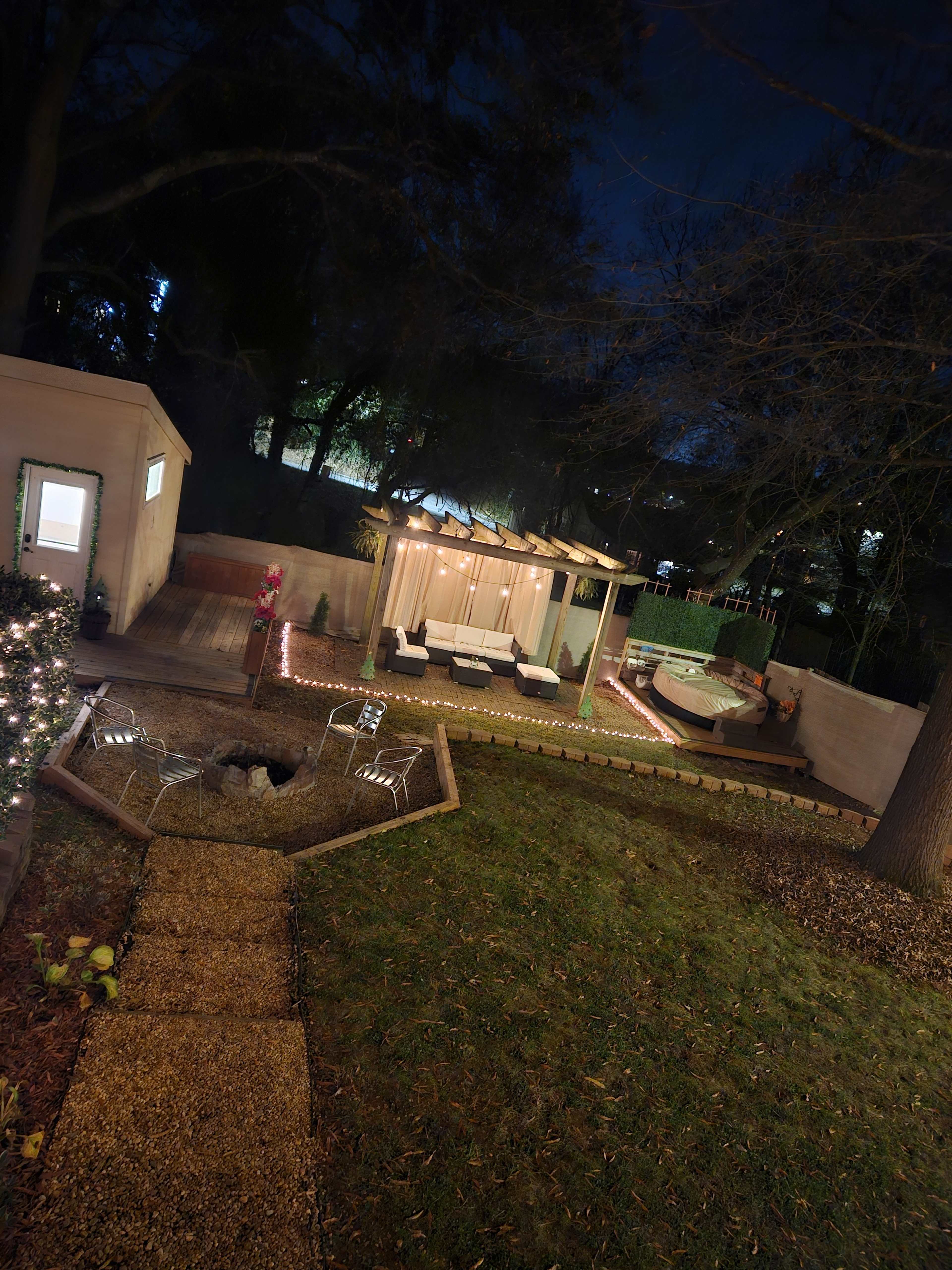 The image shows a landscaped backyard at night featuring a patio area with seating, a hot tub, and outdoor lighting.