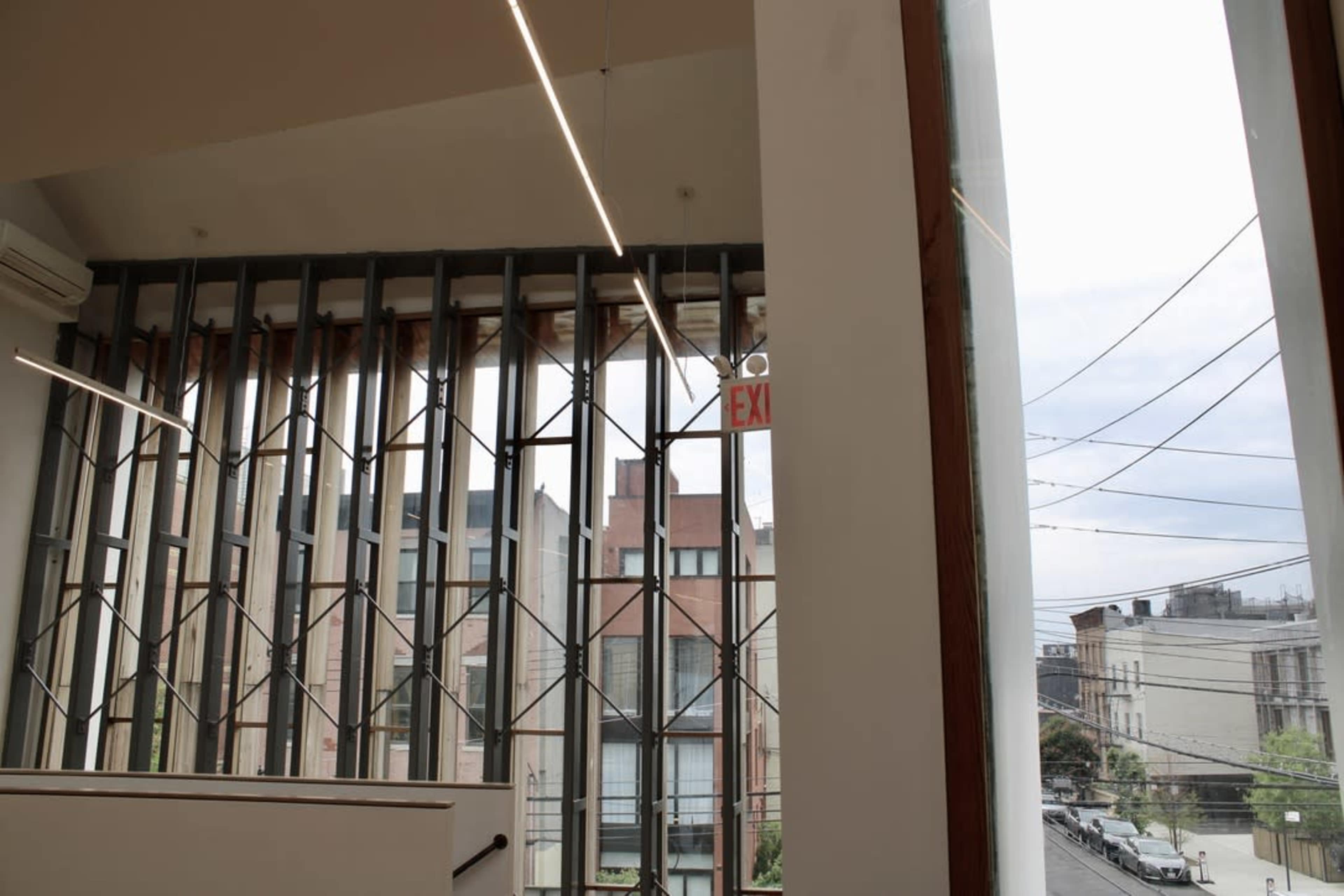 The image shows an interior view of a building with a modern design featuring a large window frame and exposed structural elements, looking out onto a street with visible power lines and buildings in the background.