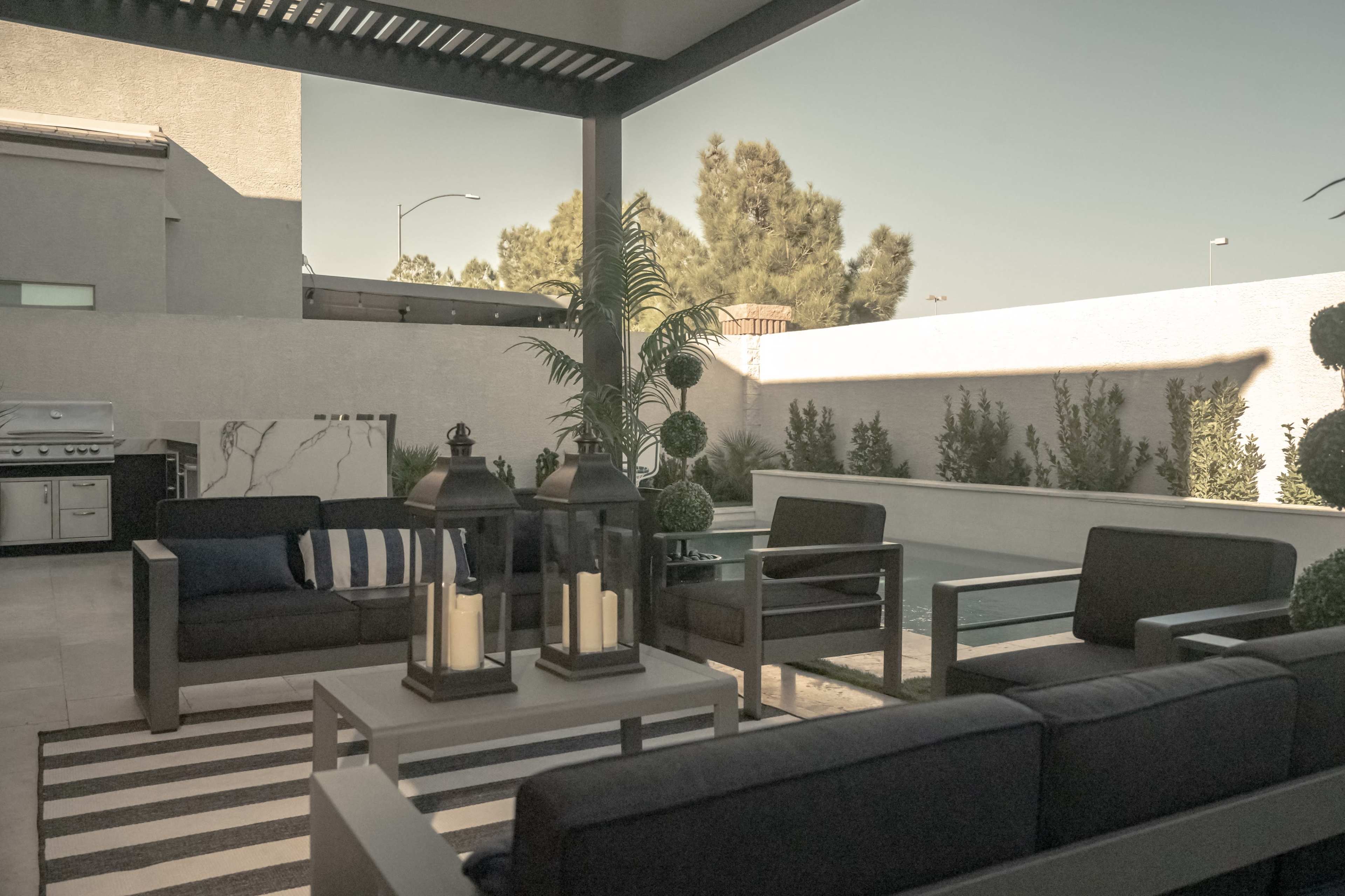 A modern outdoor patio with black seating, a striped rug, and potted plants under a shaded pergola.