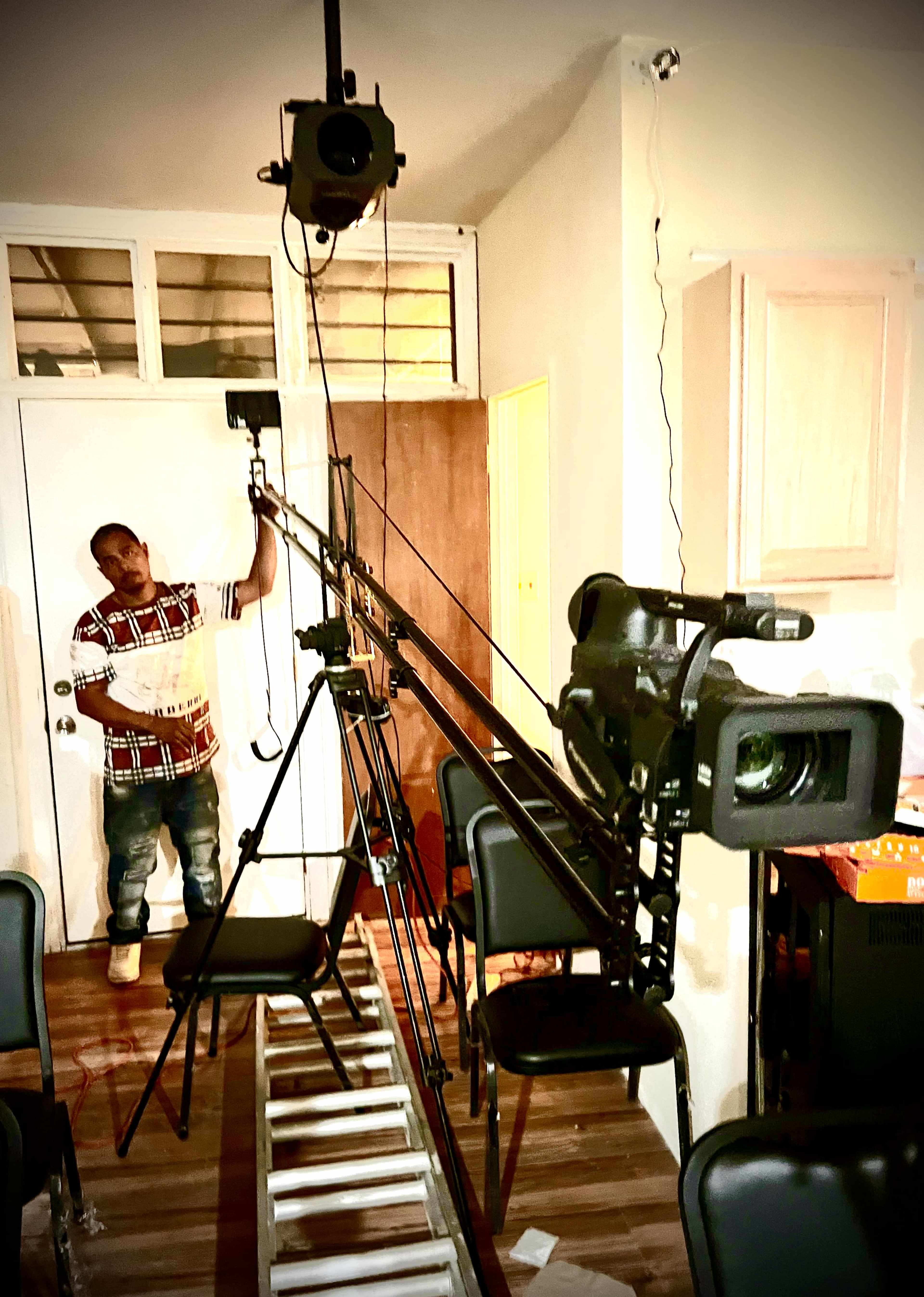 A man holds a clipboard while standing next to a camera setup with tripods and lighting in a room filled with chairs.