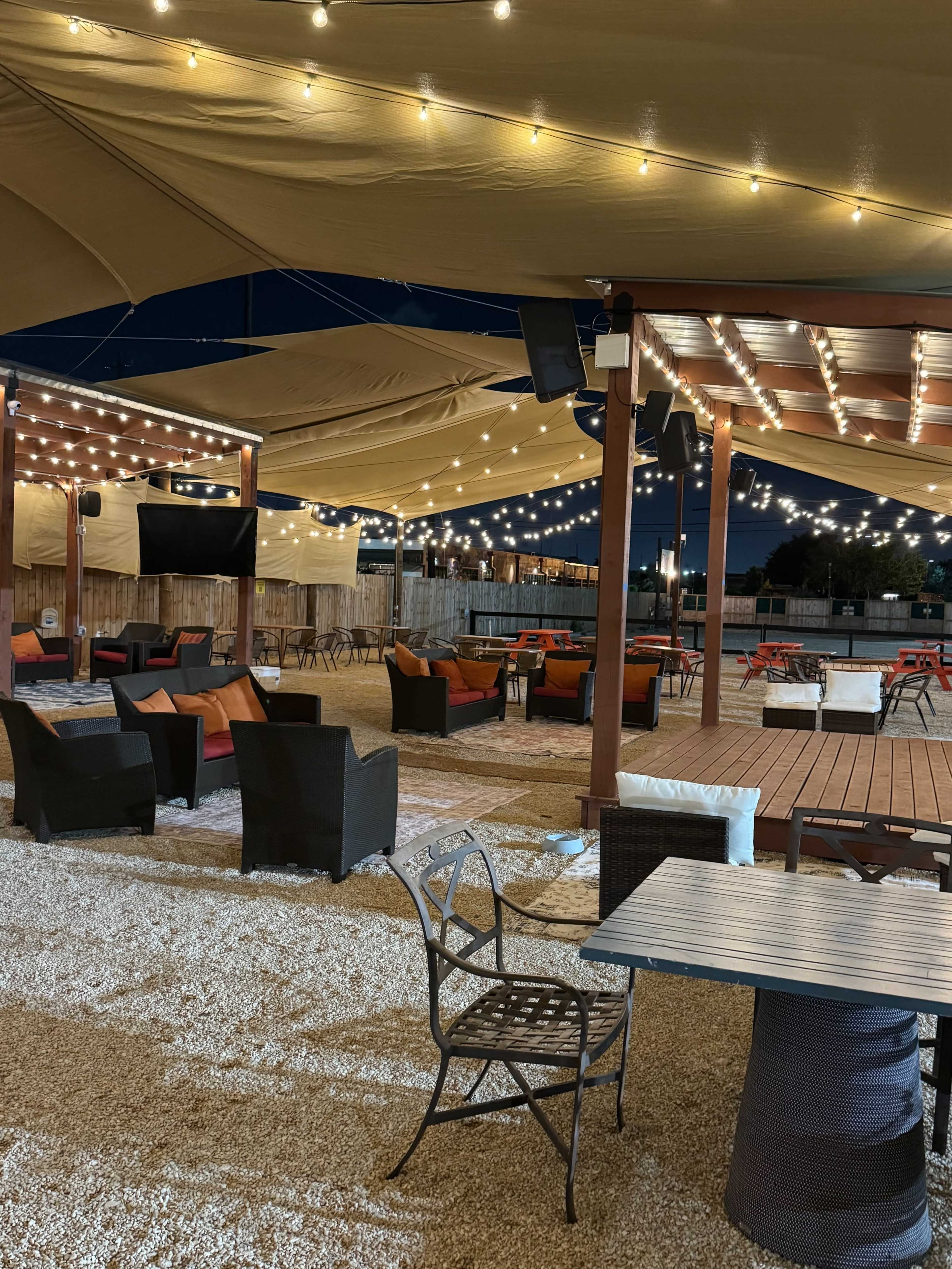 The image shows an outdoor seating area at night, decorated with string lights and featuring various tables and chairs under a large canopy.