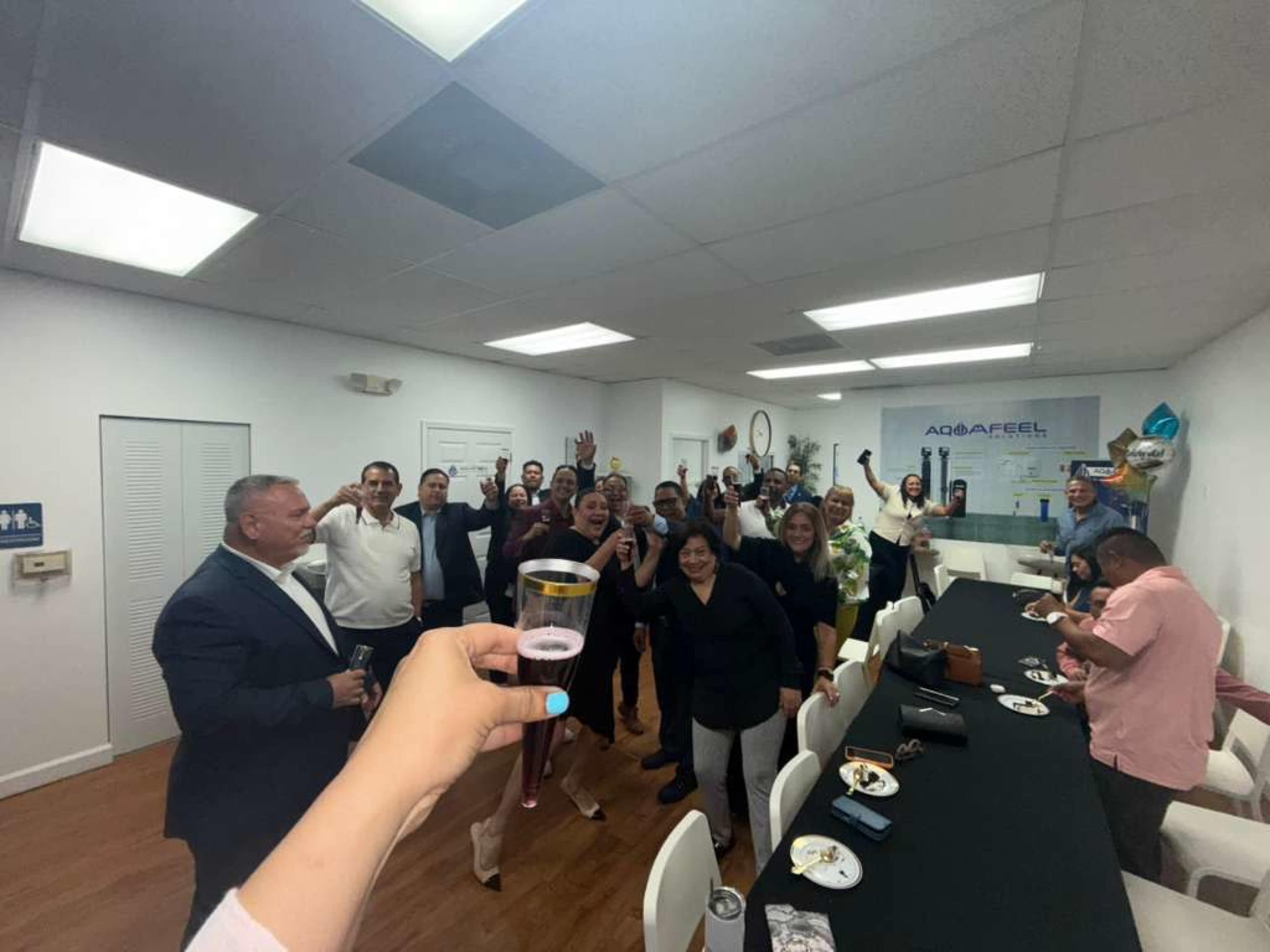 A group of people is gathered in a well-lit room, celebrating with drinks and food while some are waving and smiling at the camera.