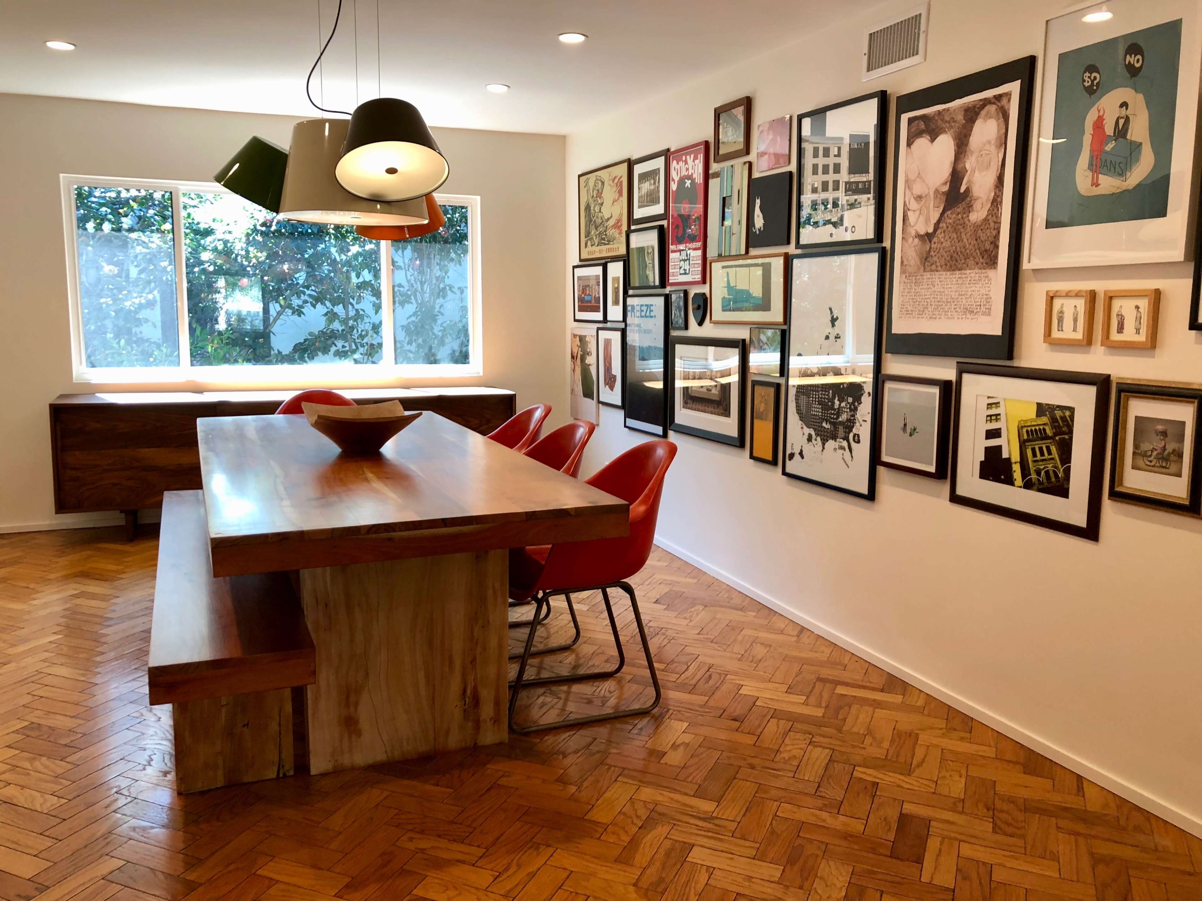 The image shows a dining area with a large wooden table and red chairs, surrounded by a wall adorned with various framed artwork.