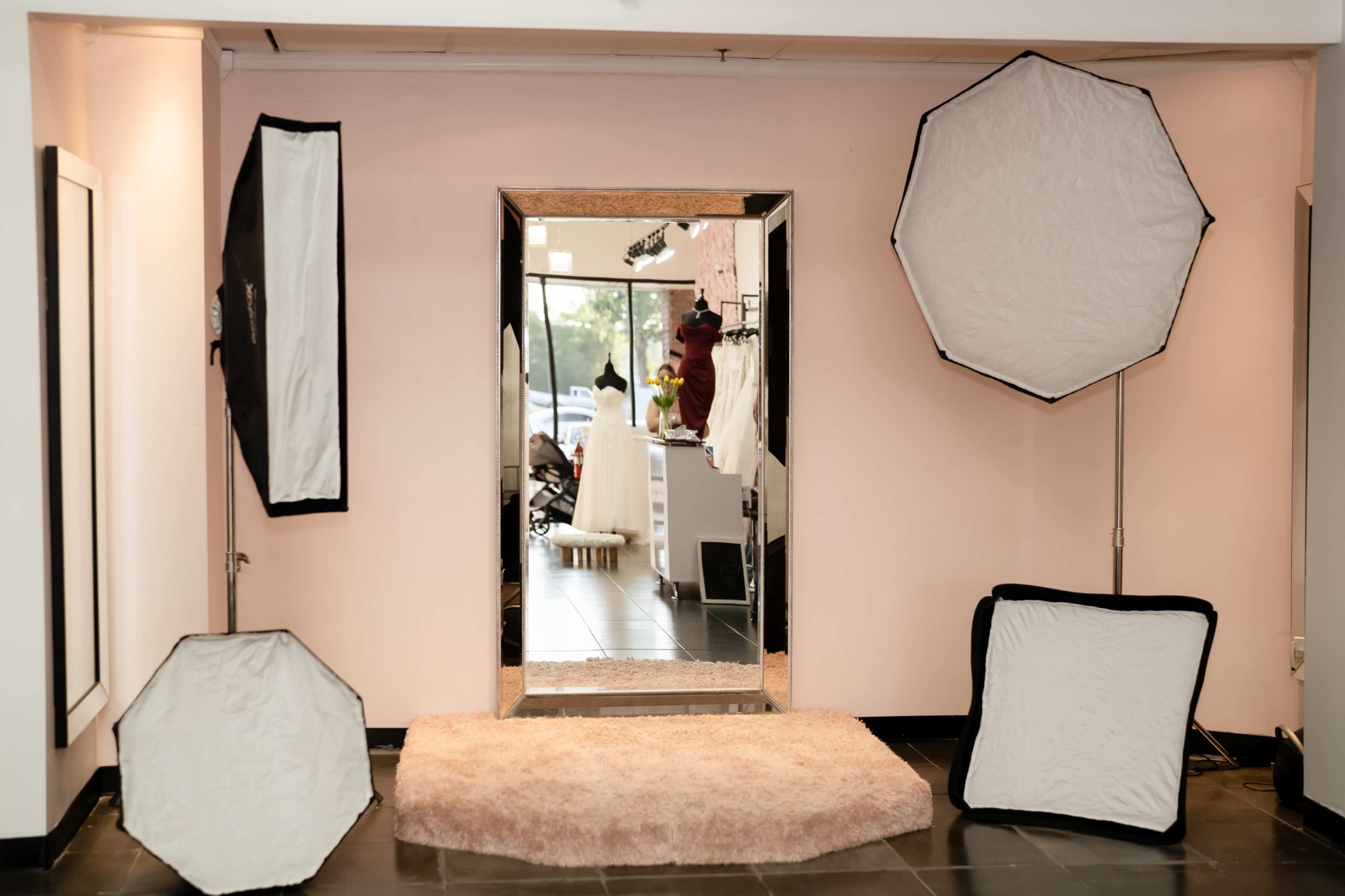A bright, modern fitting room with a large mirror surrounded by soft light reflectors on either side, set against pink walls and a black tiled floor.