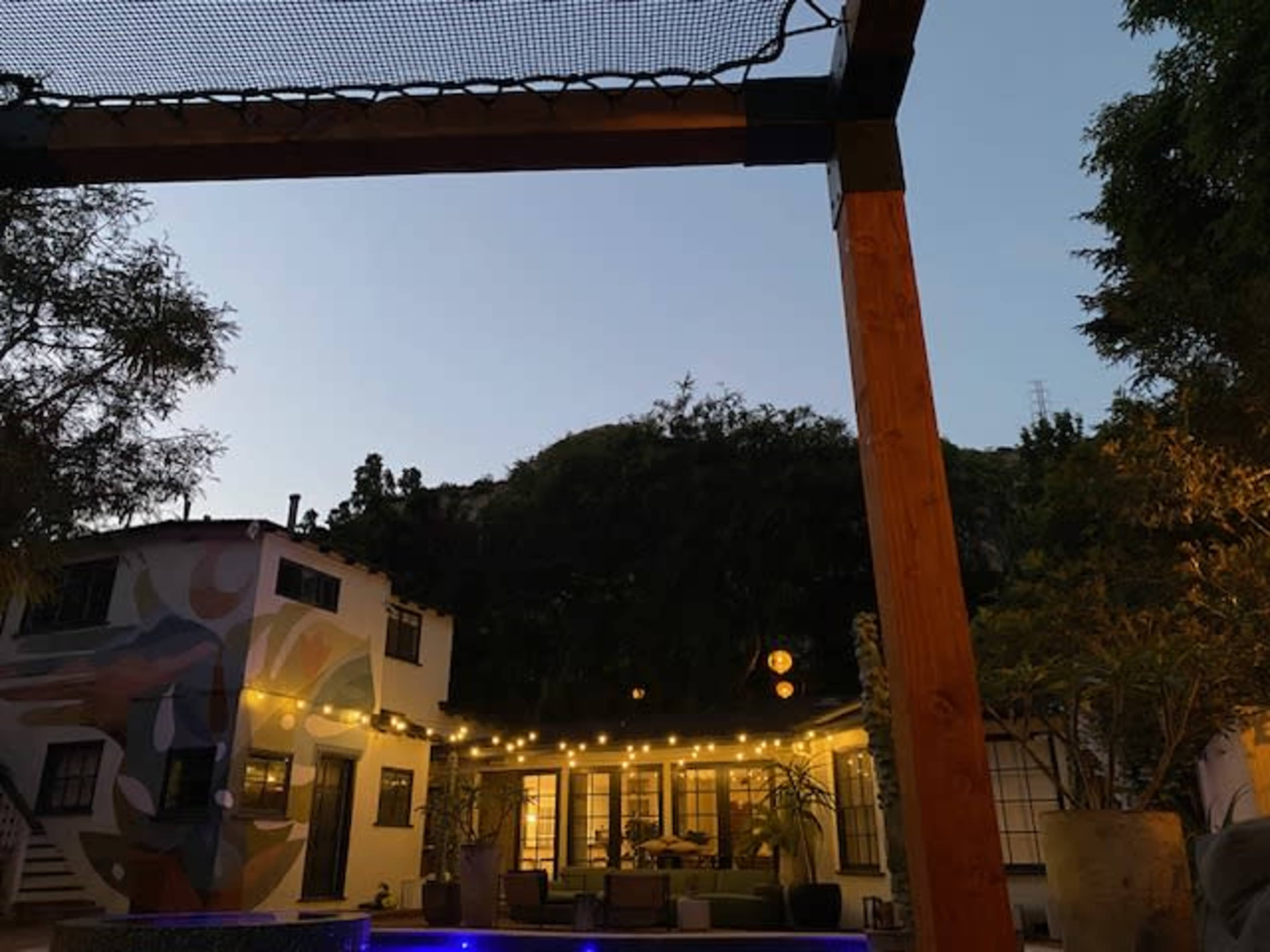 The image shows a courtyard with string lights, surrounded by buildings and greenery, under a twilight sky.