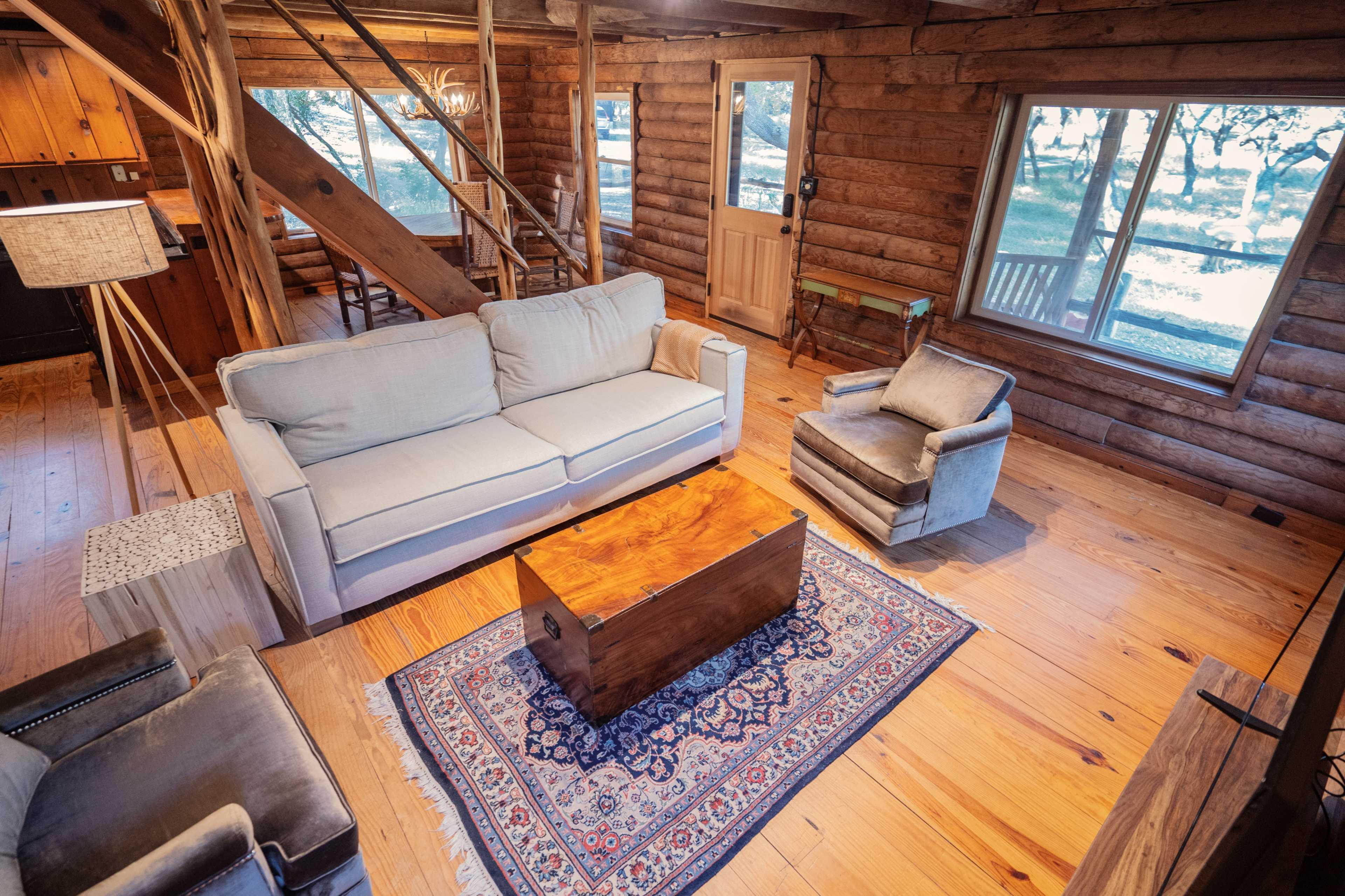 A wooden cabin interior featuring a light-colored sofa, a wooden coffee table, and a chair, with large windows offering a view of the outdoors.