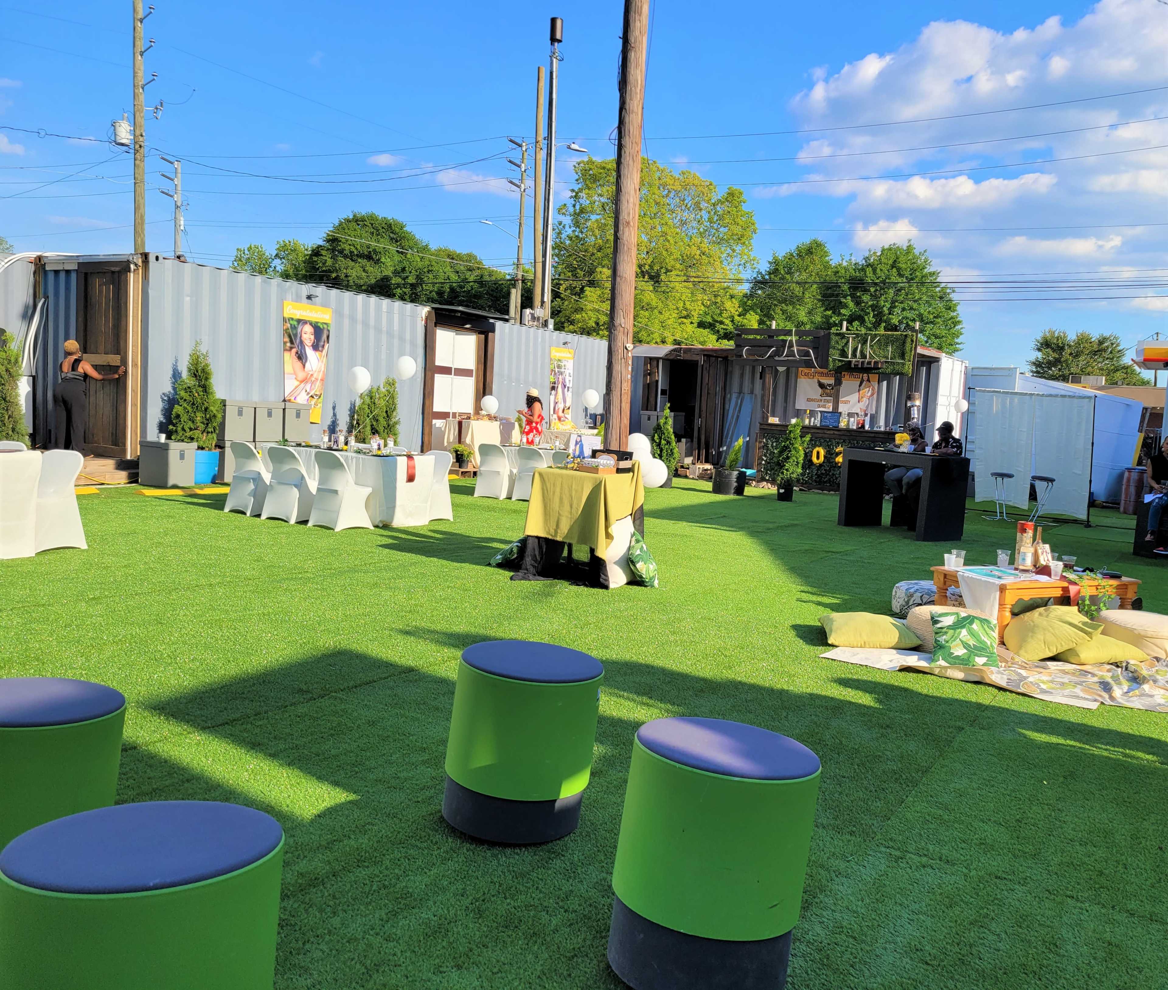 An outdoor event space with green artificial turf, seating arrangements, and shipping containers in the background.