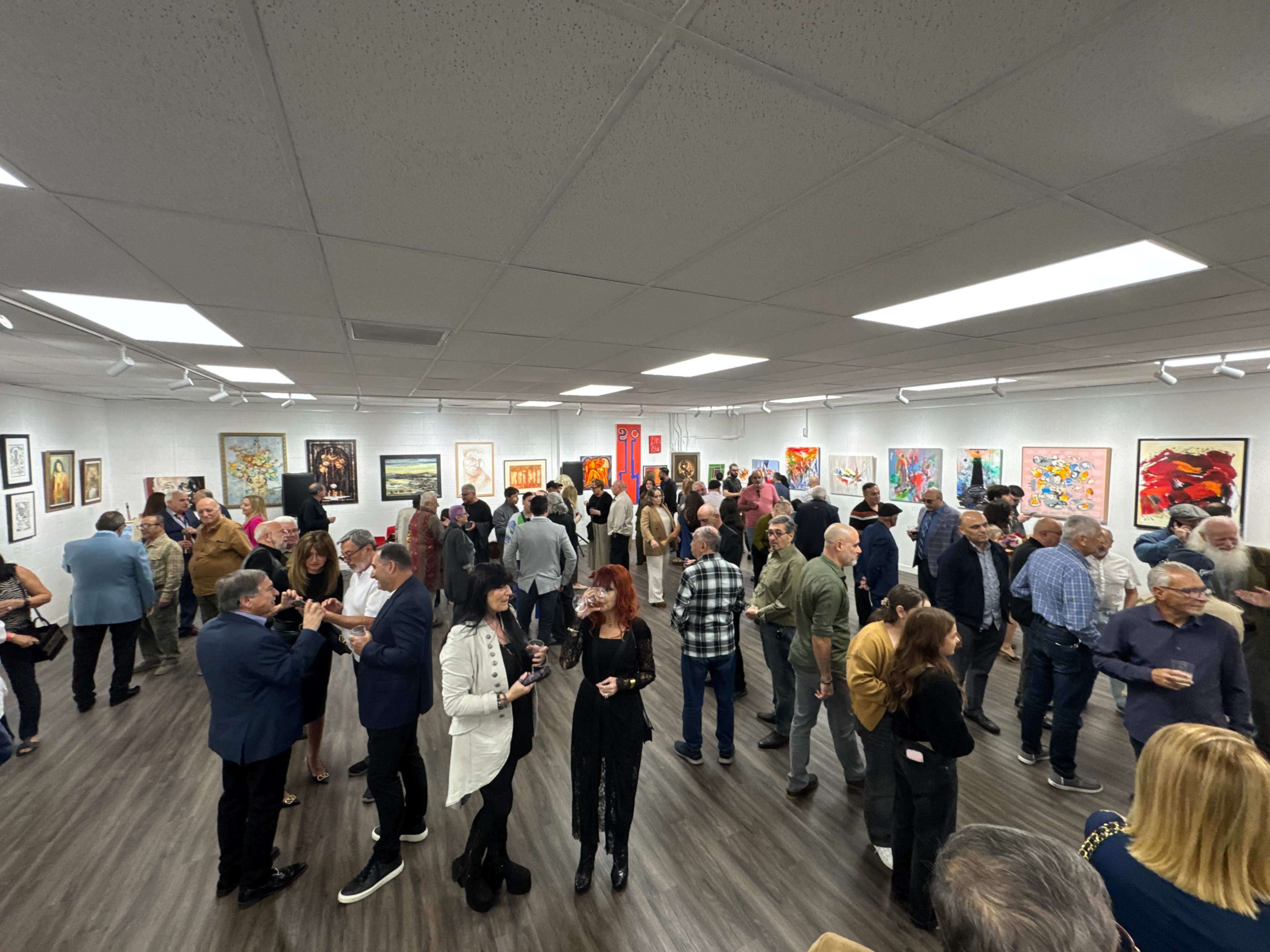 A large group of people mingles and engages in conversation at an art gallery opening, surrounded by various pieces of artwork displayed on the walls.