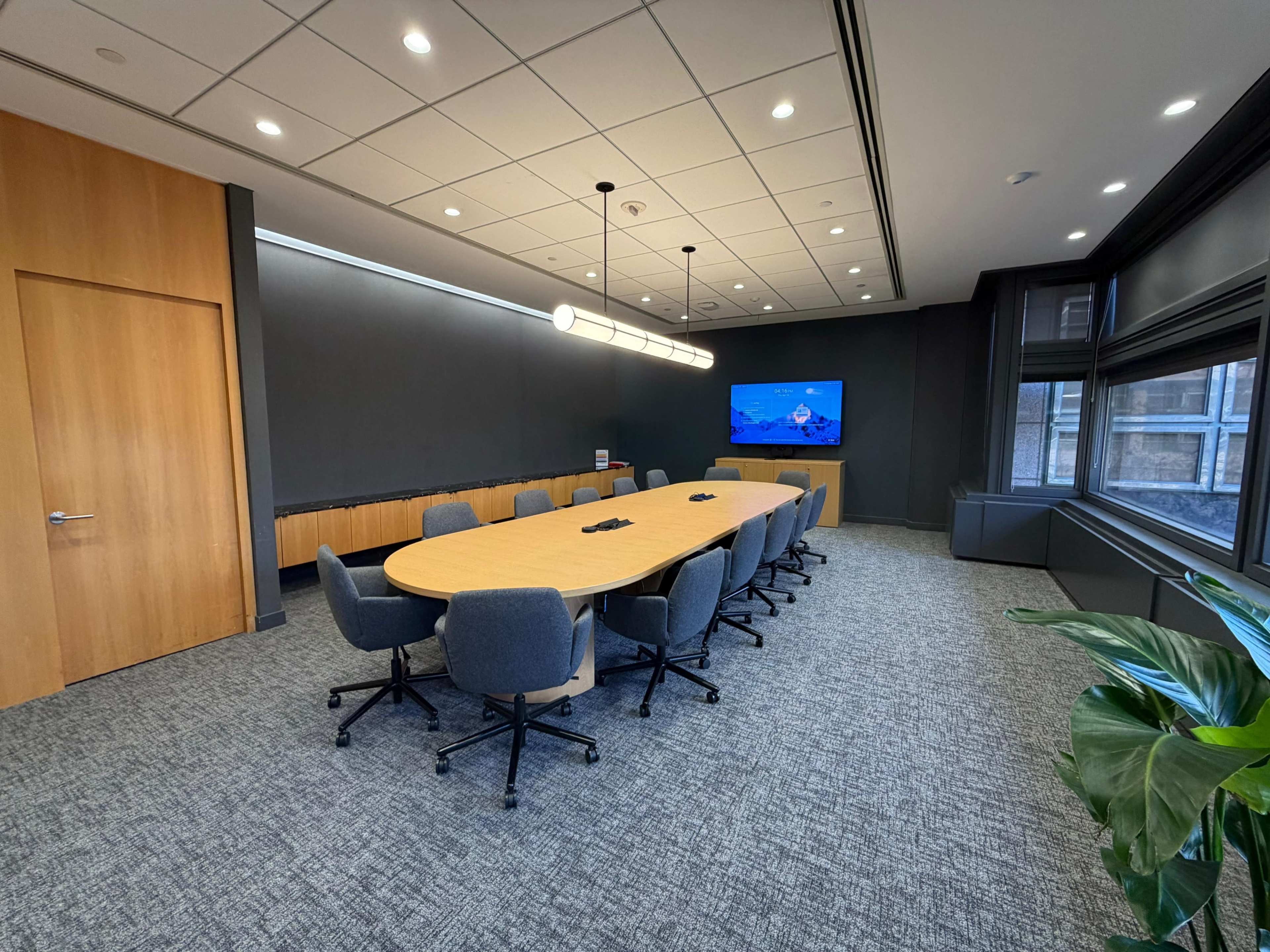 A conference room features a large oval table surrounded by upholstered chairs, with a wall-mounted screen and large windows letting in natural light.
